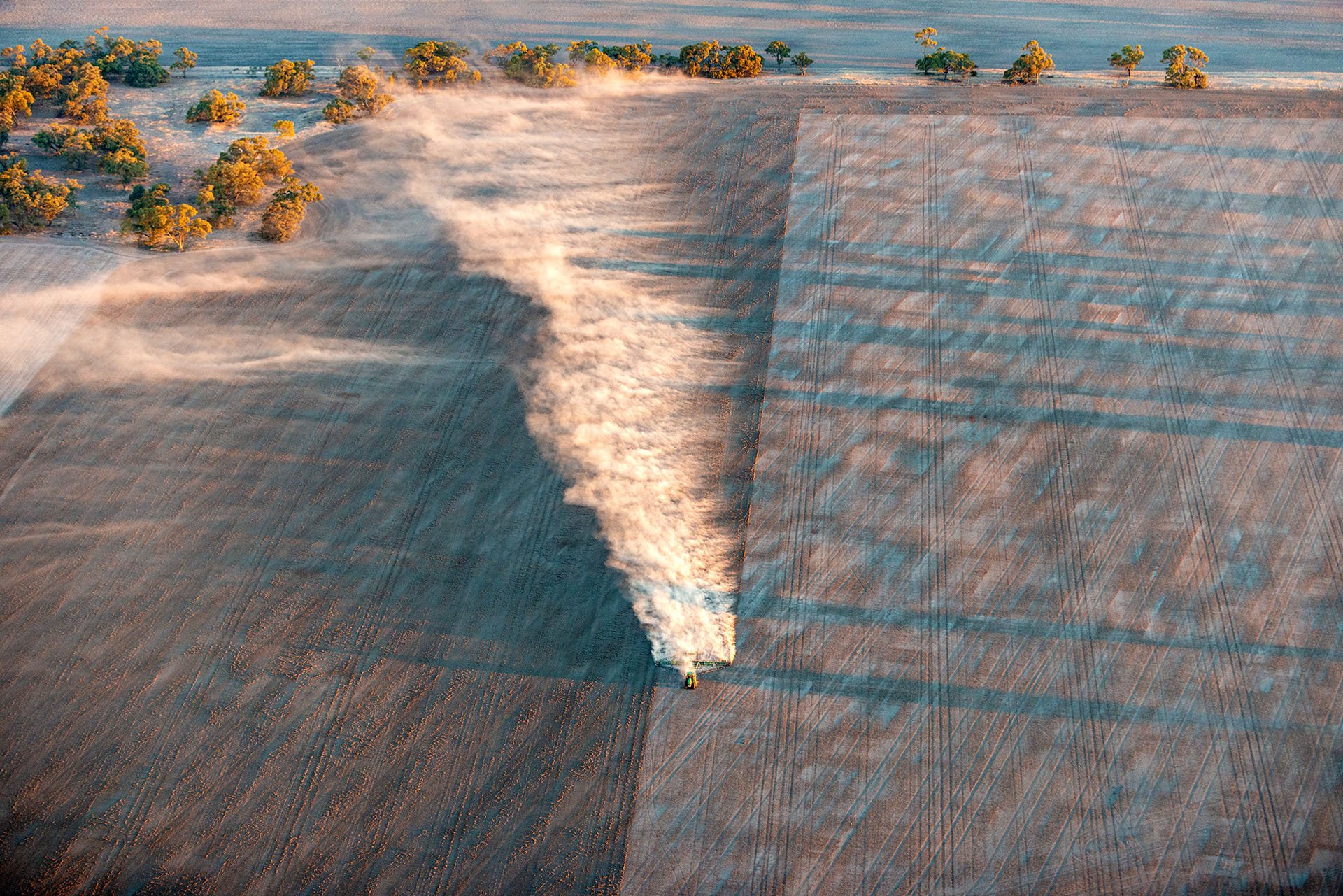 Aerial view of a tractor kicking up a long plume of dust as it works a paddock near Nhill in the Wimmera, Victoria, lit by low golden light.