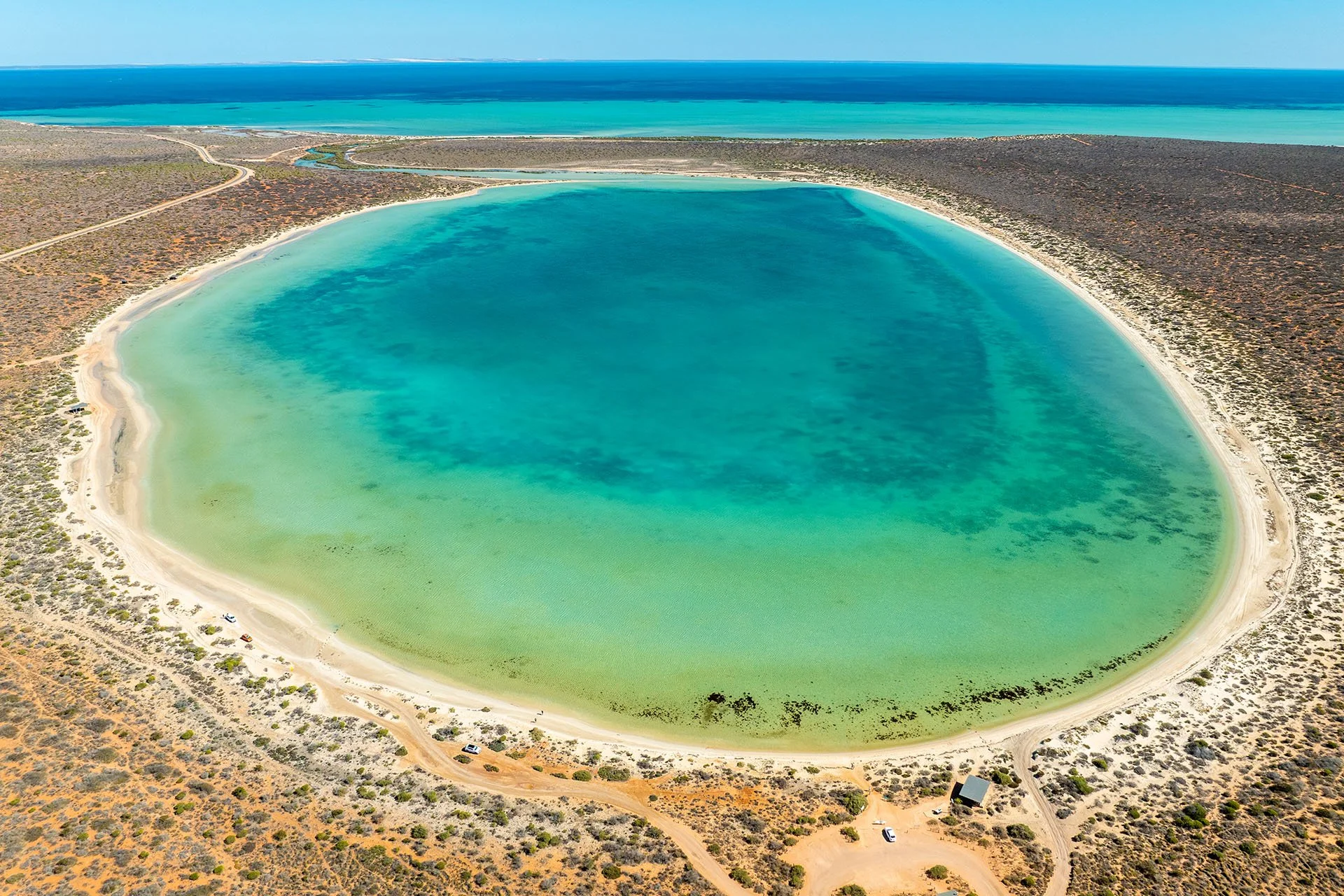 Aerial view of Little Lagoon near Denham in Shark Bay, Western Australia, a near-circular turquoise lagoon ringed by white sand and red scrubland, with the deep blue ocean beyond.