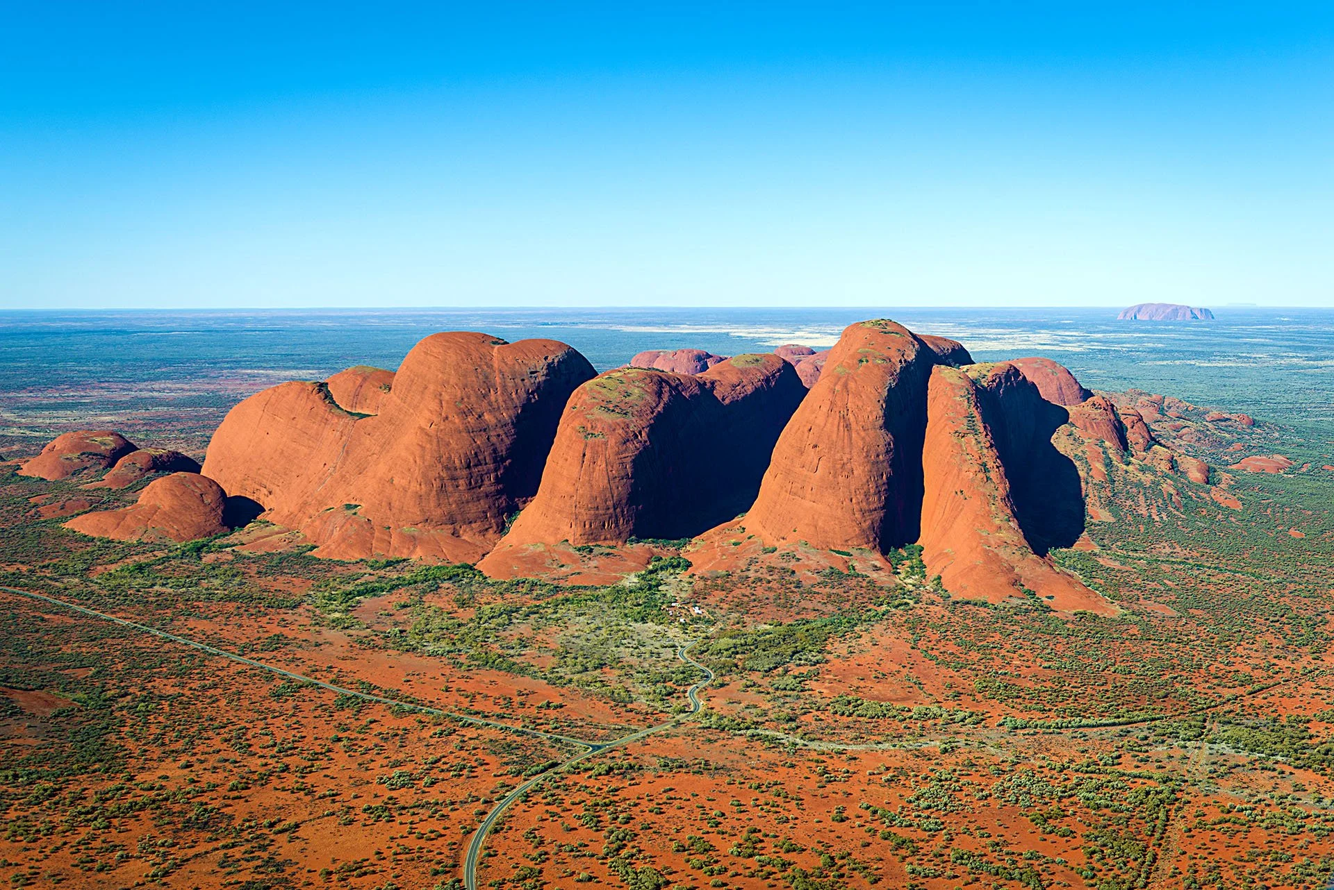 Aerial view of the red domes of Kata Tjuta rising from desert scrubland in Uluru-Kata Tjuta National Park, Northern Territory, with Uluru on the distant horizon.