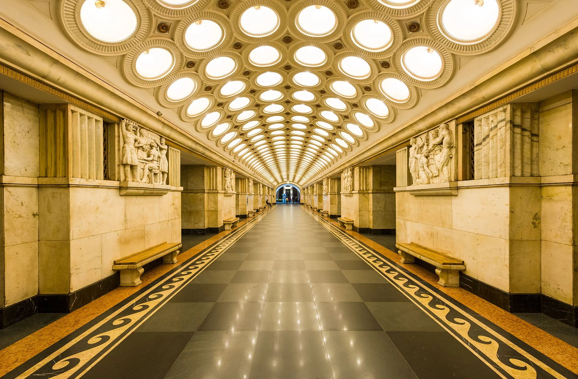 Symmetrical view along the platform of Elektrozavodskaya metro station in Moscow, Russia, with rows of round ceiling lights, marble walls, carved reliefs and a patterned floor.