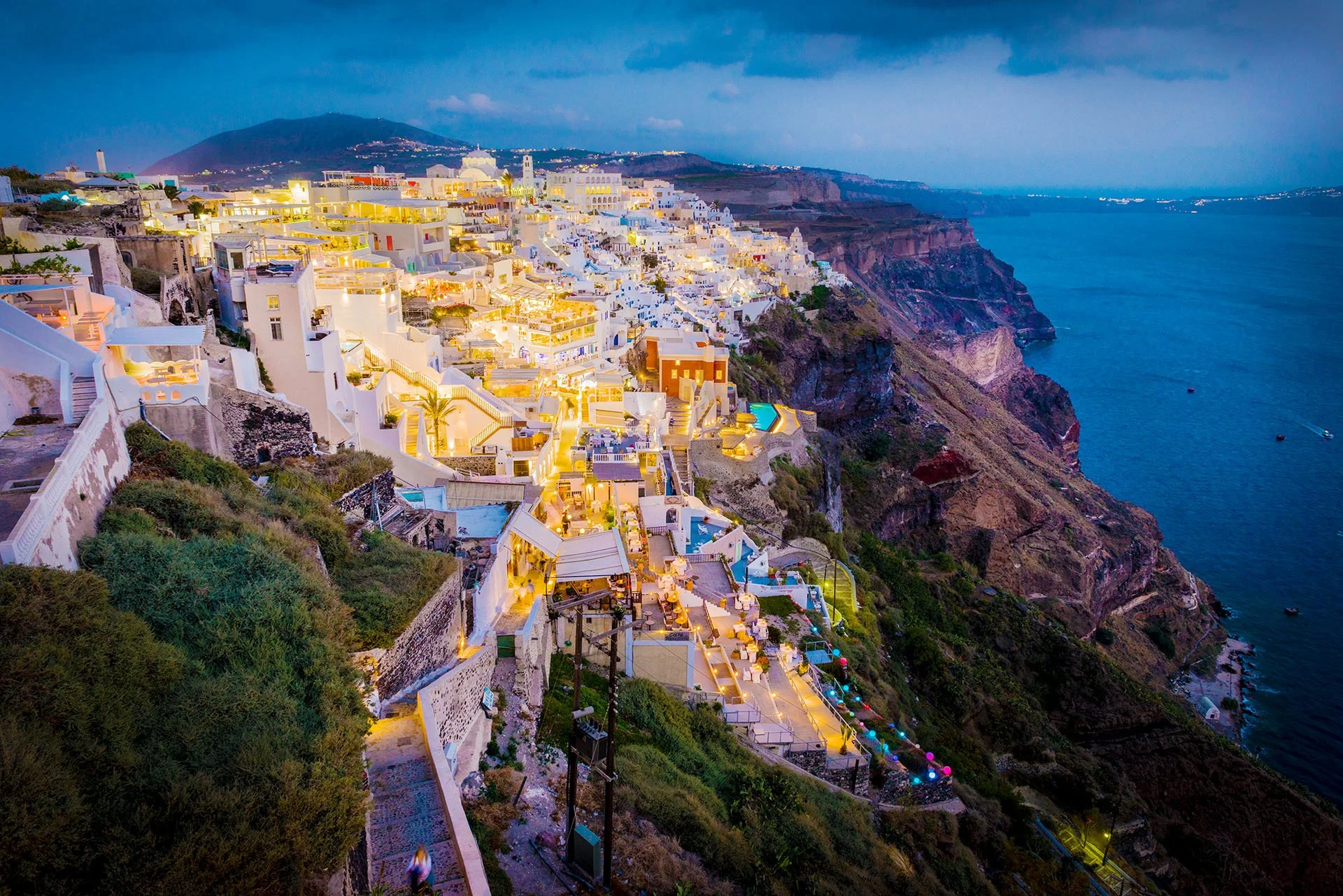 The whitewashed clifftop town of Firostefani, Santorini, Greece, glowing with warm lights at dusk above steep cliffs and the deep blue caldera.