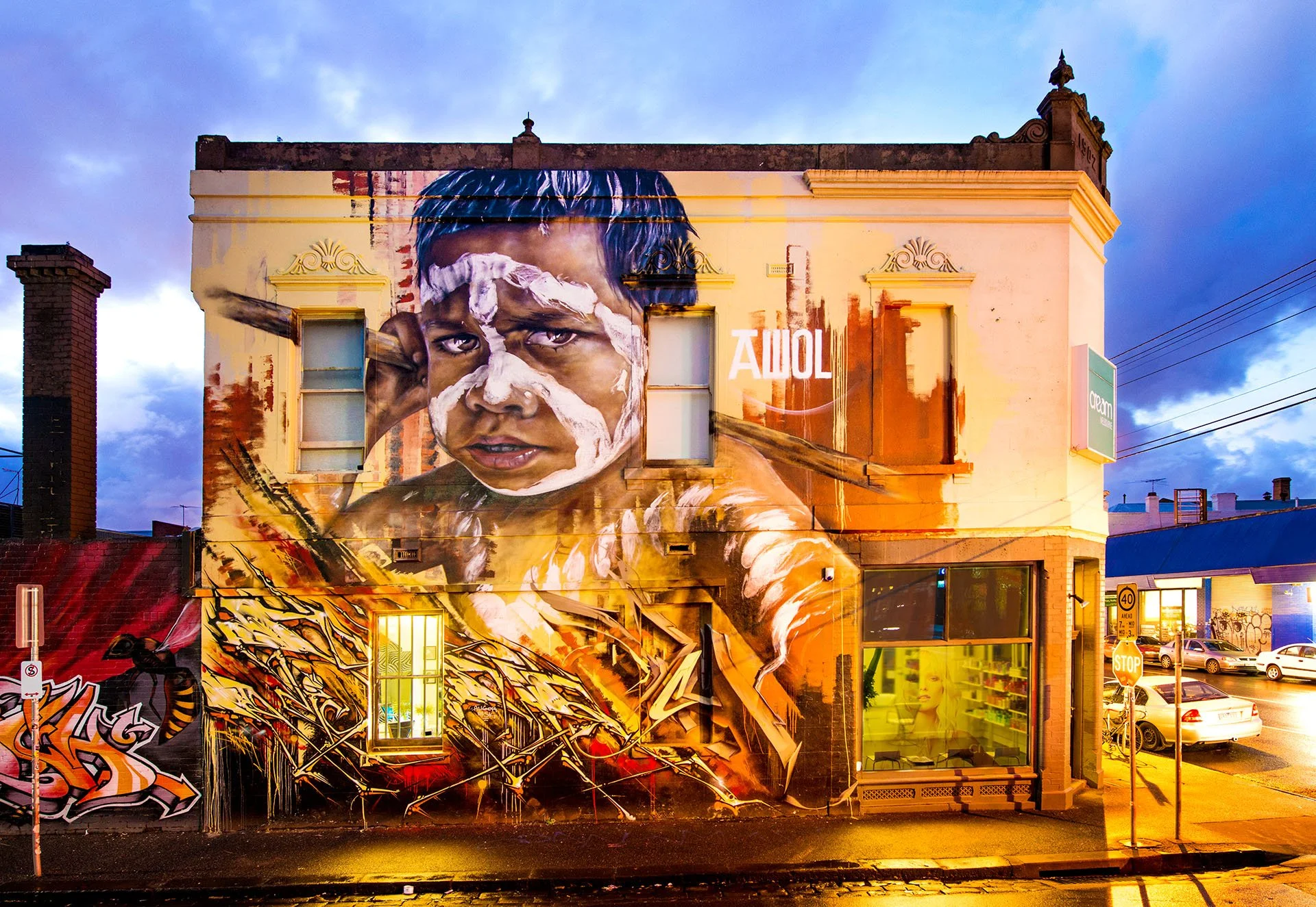Large street art mural by Adnate, Slicer and Itch on a corner building in Fitzroy, Melbourne, showing an Aboriginal boy with white ochre face paint, glowing under evening streetlights and a stormy blu