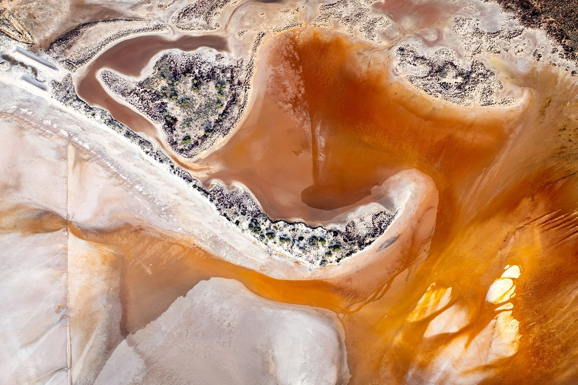 Abstract drone view of Lake Tyrrell near Sea Lake, Victoria, with swirls of orange and rust-coloured water winding around white salt flats and small scrub-covered islands.