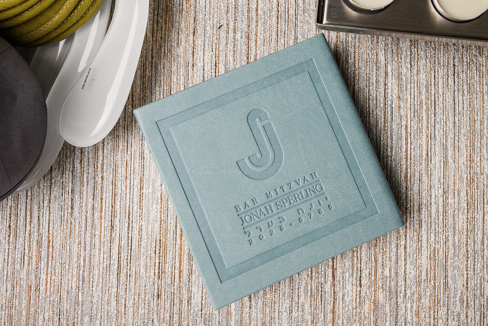A light blue box with embossed text reading 'Bar Mitzvah Jonah Sperling 5 July 2021 628.5786' on a wooden surface, next to a white dish soap bottle and part of a black and gray object.