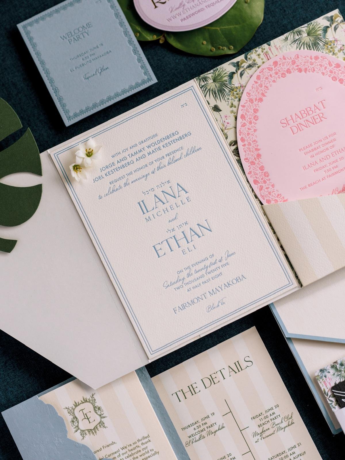 Elegant wedding invitation suite featuring a main card with blue text announcing the marriage of Ilana Michelle and Ethan Eli on June 25 at Fairmont Mayakoba, with additional pink and floral-themed event cards for Shabbat dinner and other celebrations, all decorated with small flowers and greenery.