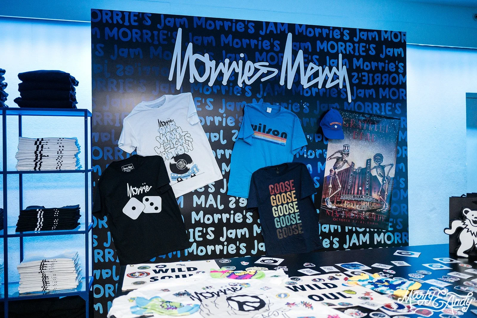 Display of Morrie's Jam merchandise including T-shirts with various designs, a cap, and a poster, set against a blue backdrop with the Morrie's Jam logo.