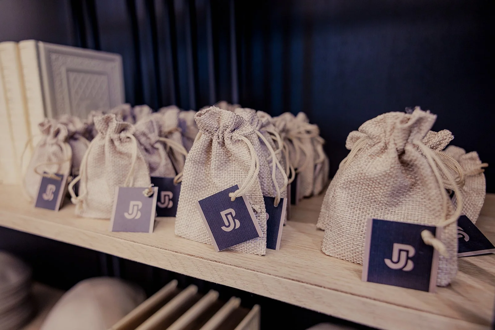 Small beige drawstring pouches with tags featuring a stylized 'S' logo, arranged on a wooden shelf.