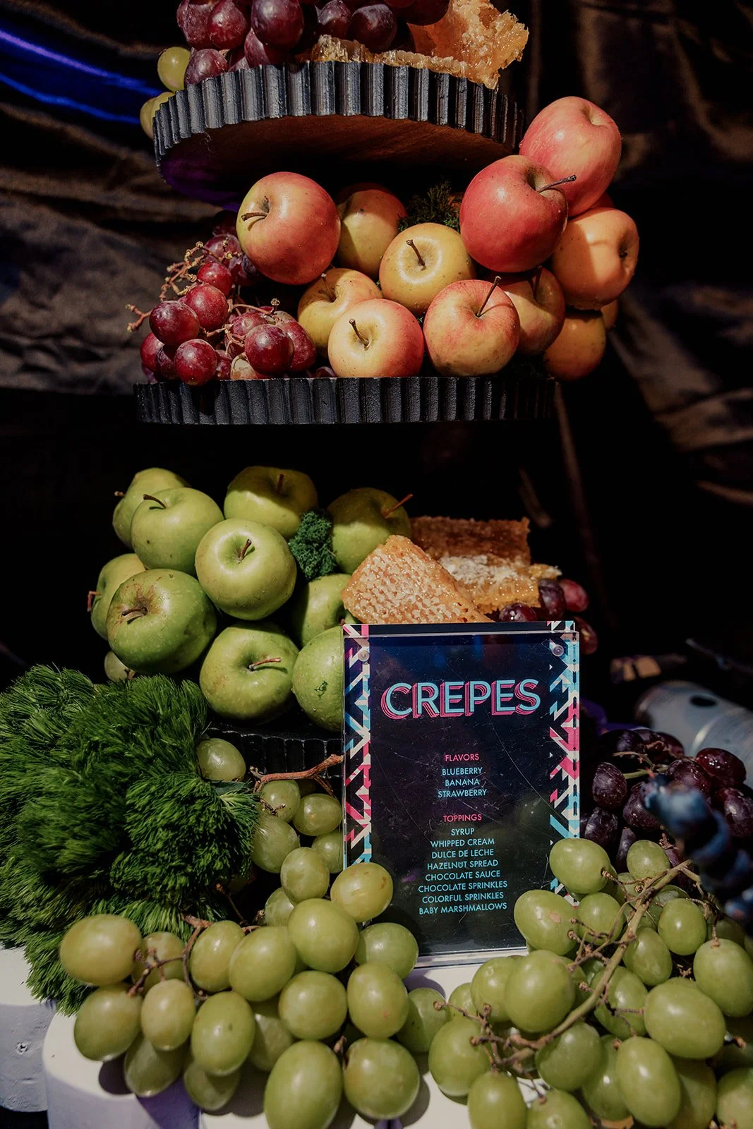 A display of fresh green and red apples, grapes, and honeycomb with a sign advertising crepes with blueberry, banana, and strawberry flavors, and toppings including syrup, whipped cream, and chocolate.