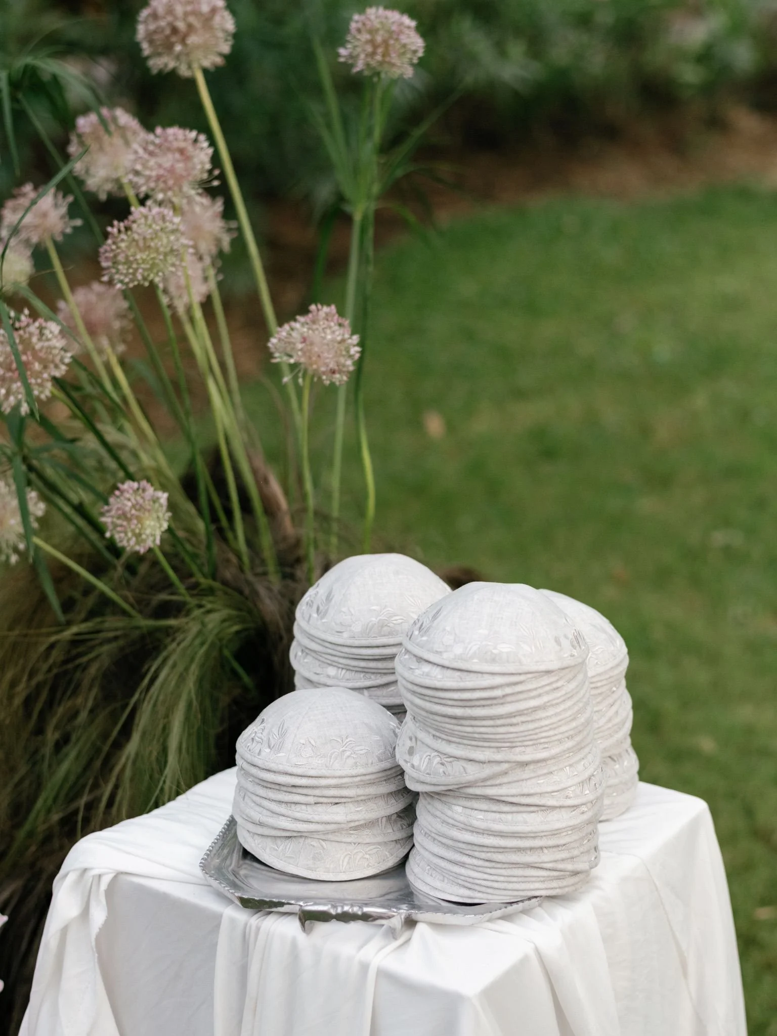 Stacked white disposable bowls with intricate floral patterns on a silver tray, placed on a table covered with a white cloth, outdoors with green grass and pinkish flowers in the background.