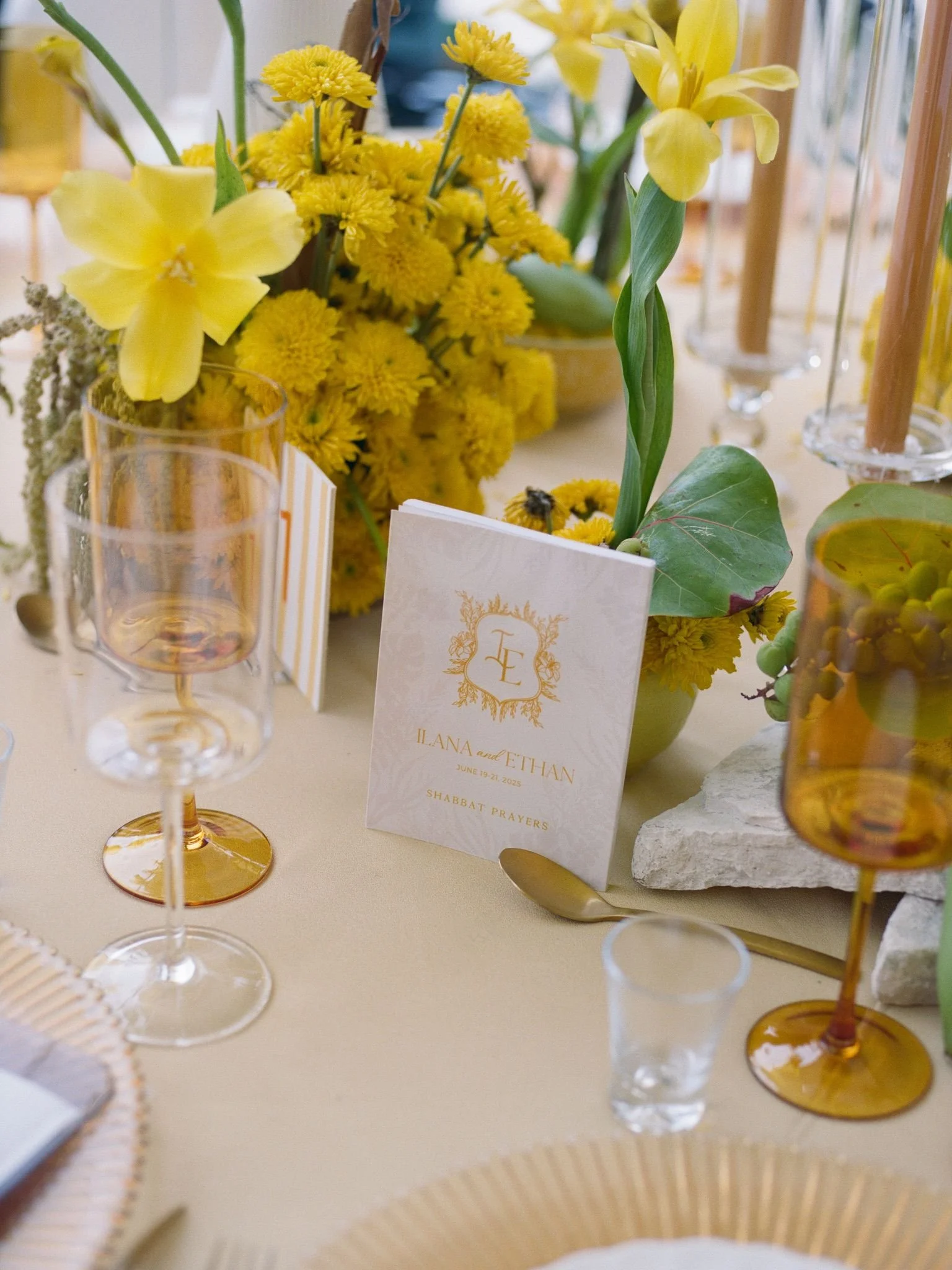 A table decorated with yellow flowers, including daisies and orchids, and surrounded by clear and amber wine glasses. There is a white card with gold and black lettering that reads 'Ilana and Ethan, June 19-21, 2025, Shabbat Prayers.'