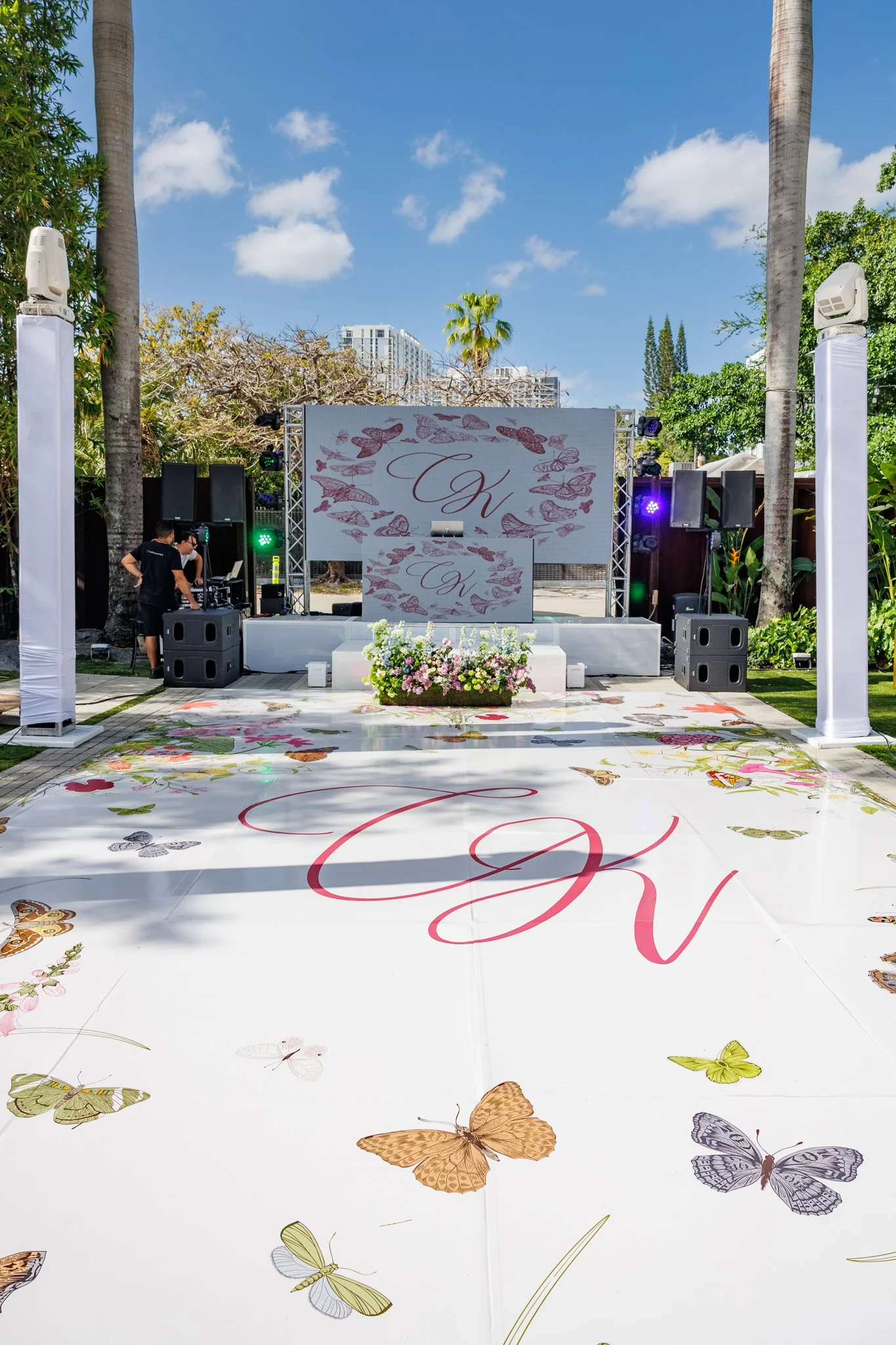 An outdoor wedding stage decorated with pink and purple butterfly motifs, and a large floral arrangement in front with pink, white, and purple flowers. There are two large screens with initials 'K' and 'V' and a DJ setup on the side. The area is surr