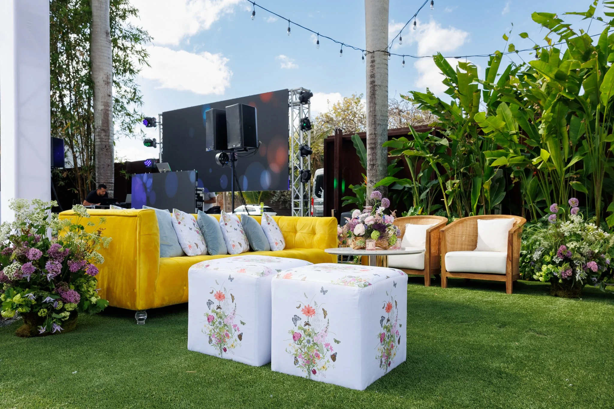 Outdoor event setup with a yellow velvet sofa, white floral-patterned table, wicker chairs, and lush greenery, including large plants and flowers. In the background, there is a stage with lighting and sound equipment under a partly cloudy sky.