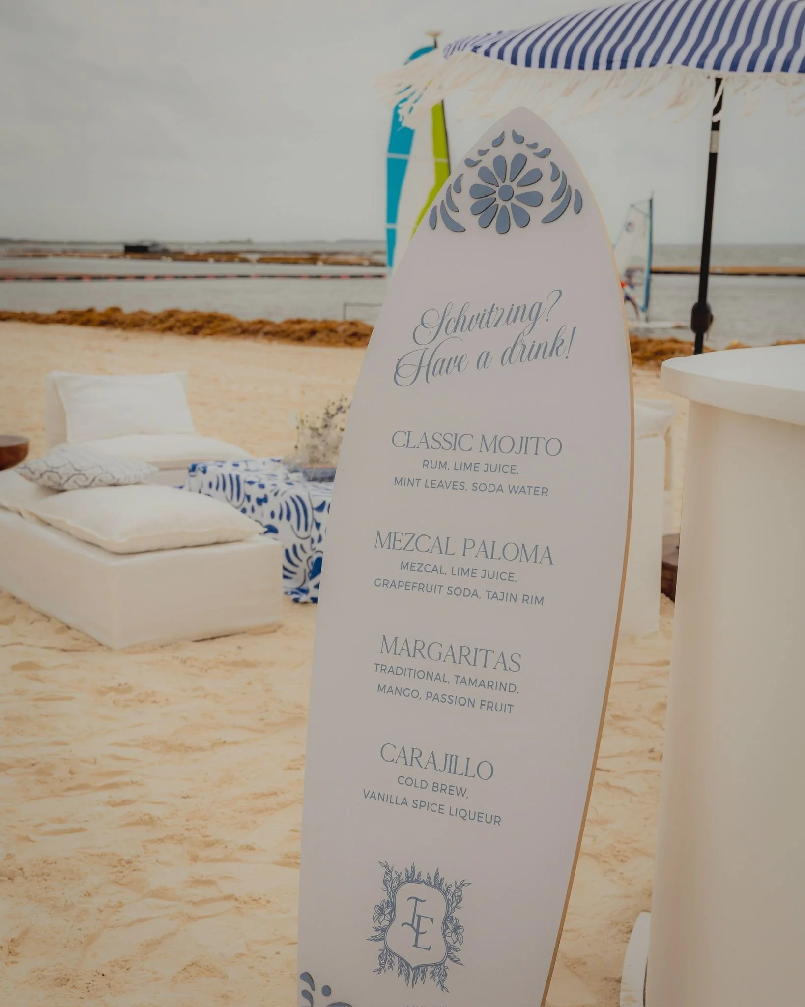 A beachside bar or drink stand with a menu sign displaying various cocktails, including Classic Mojito, Mezcal Paloma, and Carajillo. There are beach umbrellas, lounge chairs, and a sandy beach in the background.