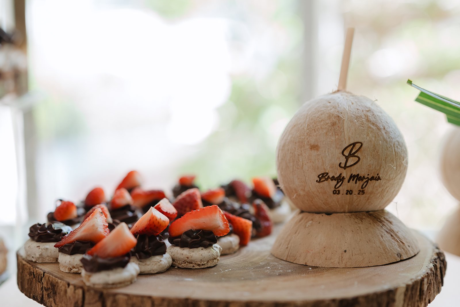 A coconut with personalized engraving, placed on a wooden base, with sliced strawberries and chocolate-covered cookies or pastries on a wooden surface in front, and a blurred background of a bright window.