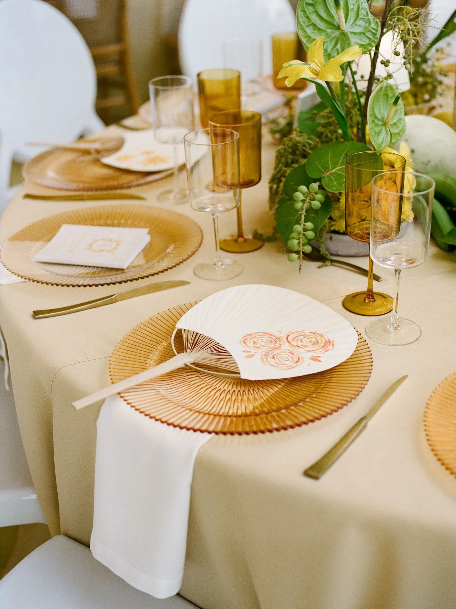 A table set for a meal with beige placemats, white napkins with a gold design, clear and amber-colored wine glasses, and a floral centerpiece with green leaves and yellow flowers.