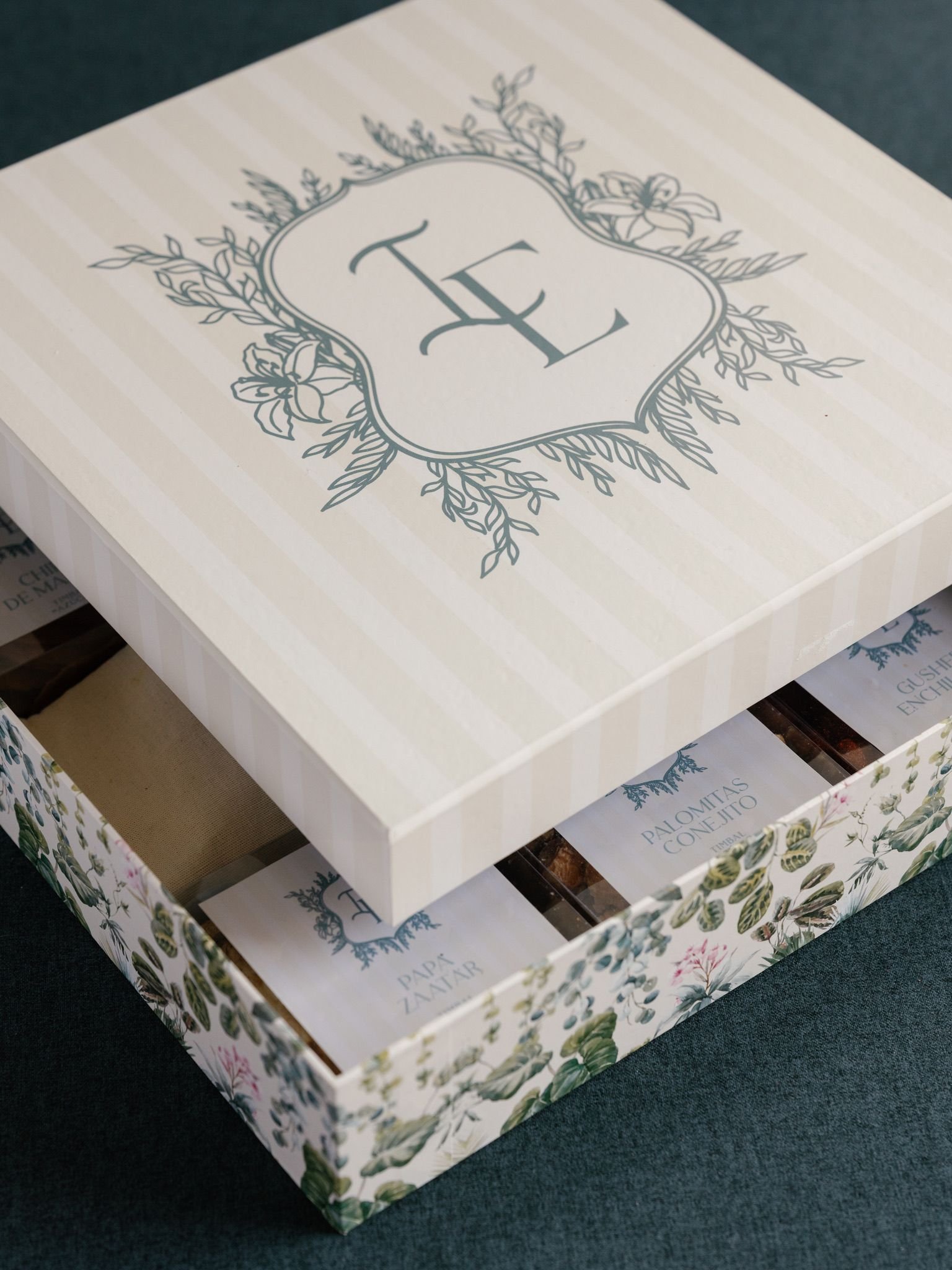 A white box with light beige vertical stripes and a floral design on the top, with the initials 'E' in the center, surrounded by greenery and flowers. Inside the box, there are various cards with floral borders and text in Spanish, including titles l