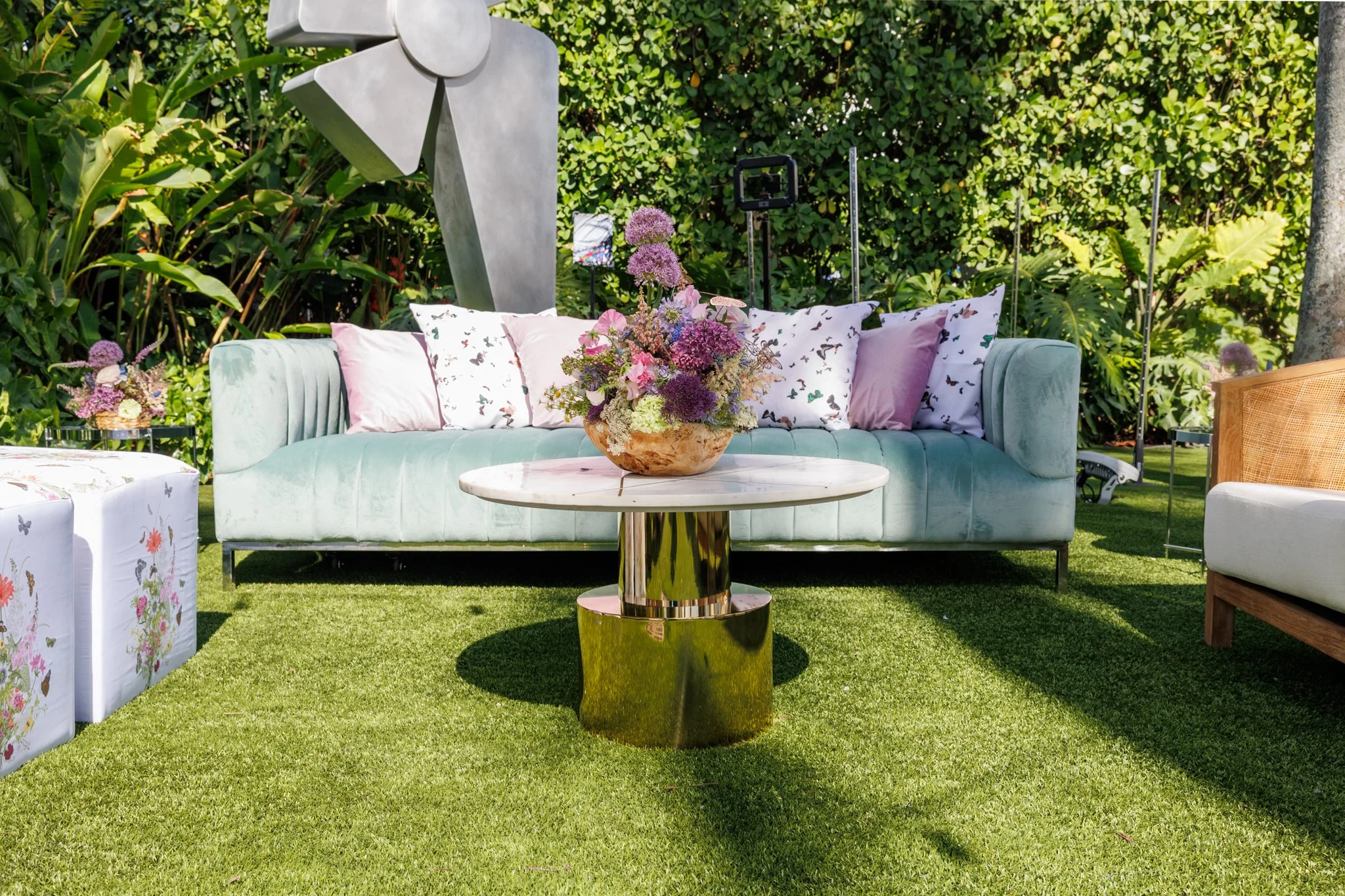 An outdoor seating area with a pastel green velvet sofa adorned with butterfly-printed pillows. In front of the sofa is a round, gold-colored coffee table with a large flower arrangement in a wooden vase. There are lush green plants and trees in the 