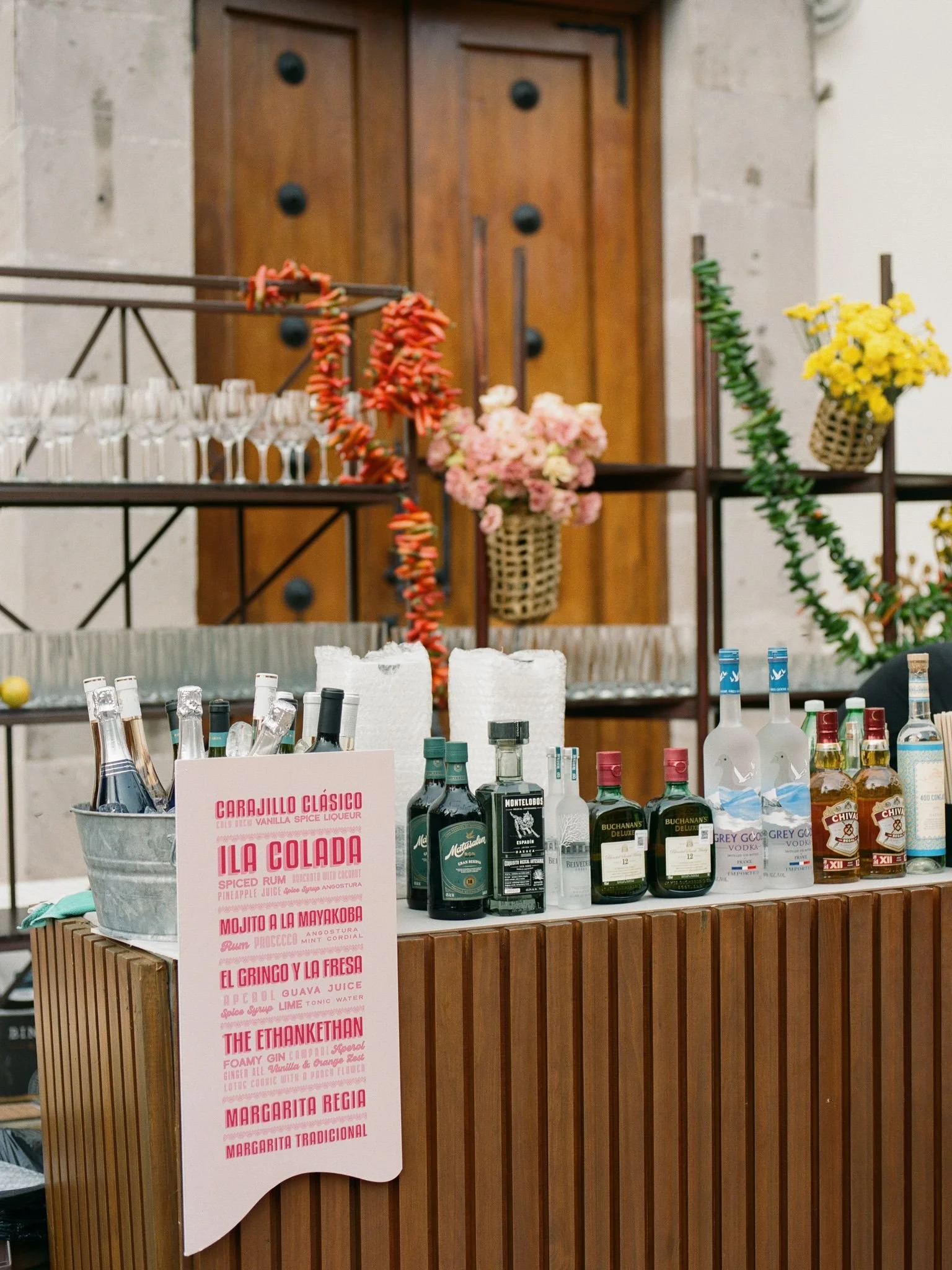A bar display with various bottles of alcohol, flowers, and a menu for cocktails on a wooden surface in front of a wooden door.
