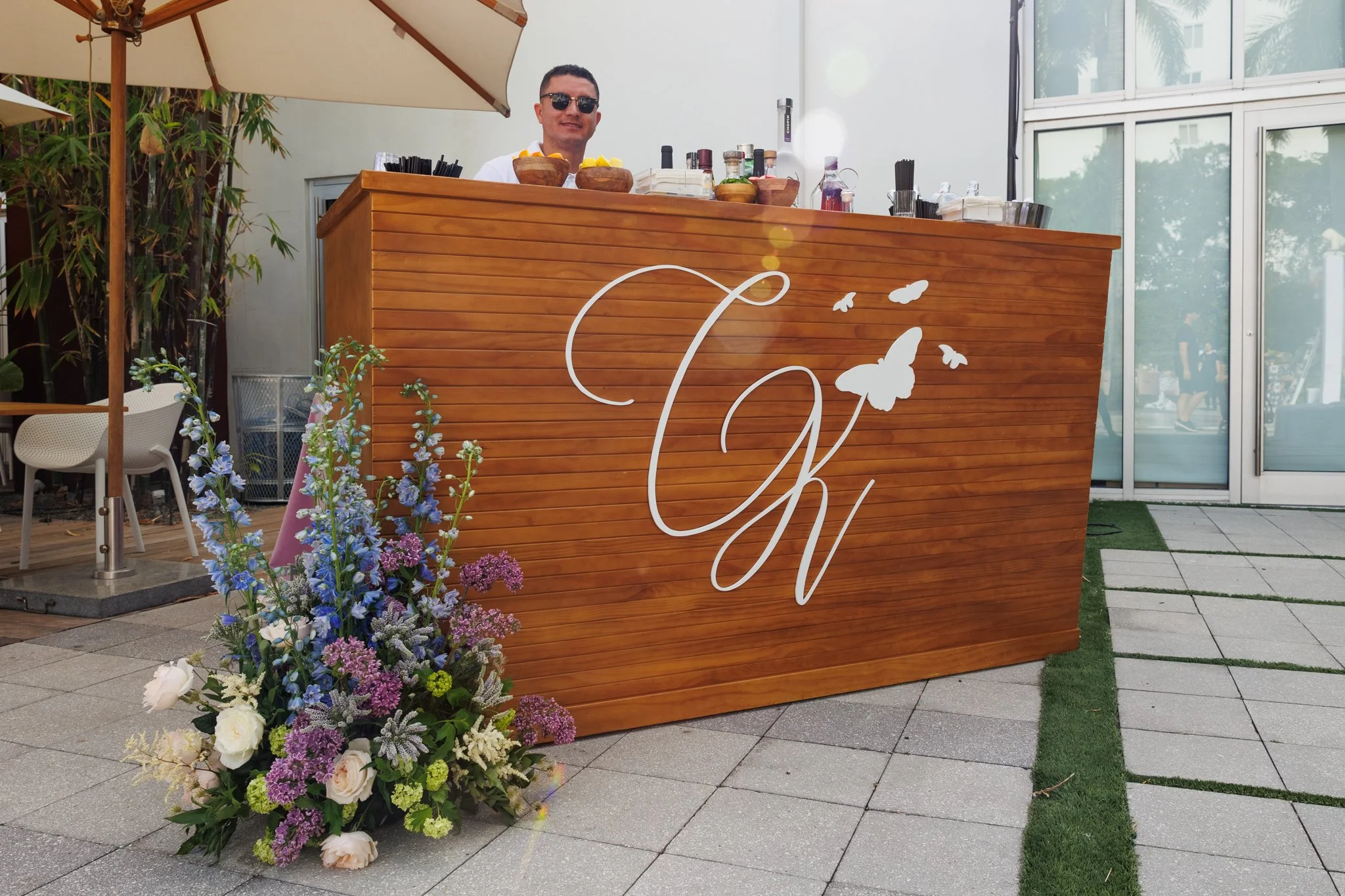 A man standing behind a wooden bar at an outdoor event, with a large floral arrangement in front of him. The bar has a white monogram with butterflies, and there are bottles, bowls, and utensils on top. Large windows and umbrellas are in the backgrou
