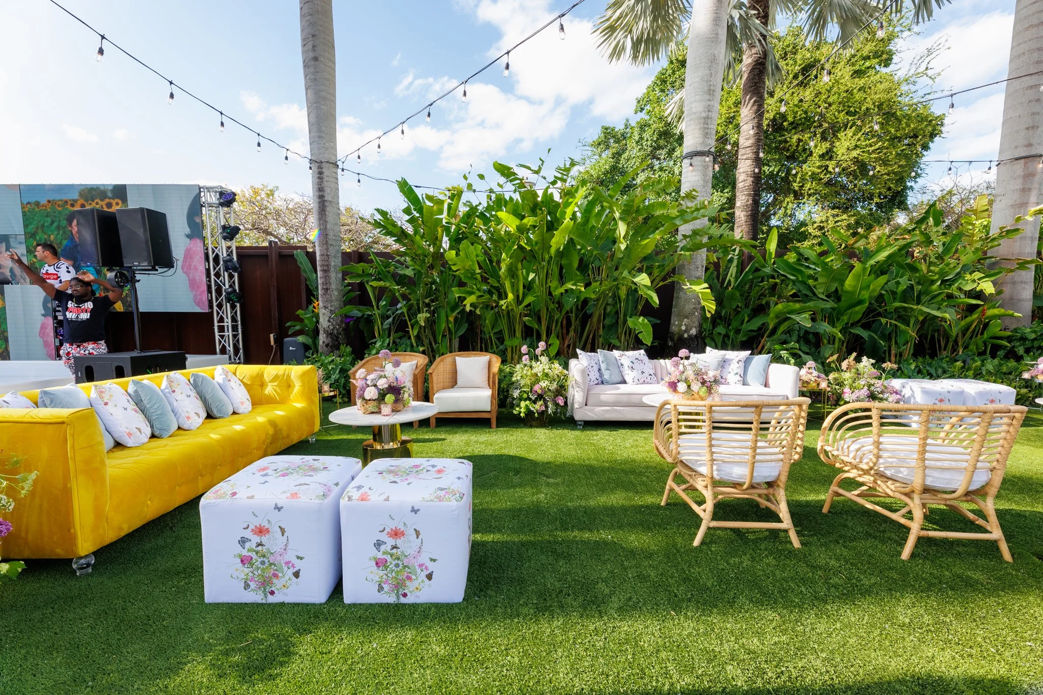 Outdoor event space with colorful furniture, floral arrangements, and string lights, surrounded by lush greenery and tall palm trees.