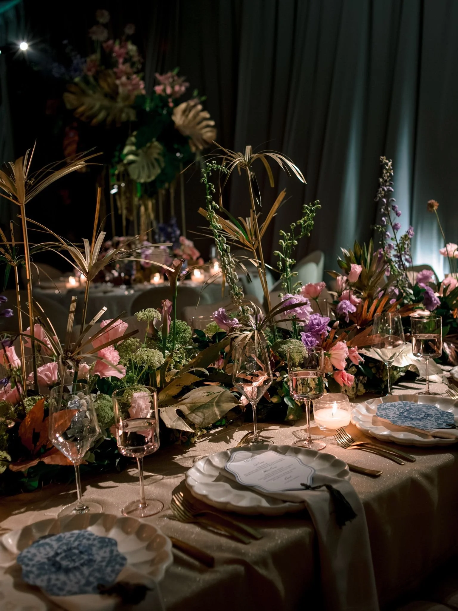 Elegant dinner table decorated with flowers, glassware, and plates in a dimly-lit setting with dark curtains in the background.
