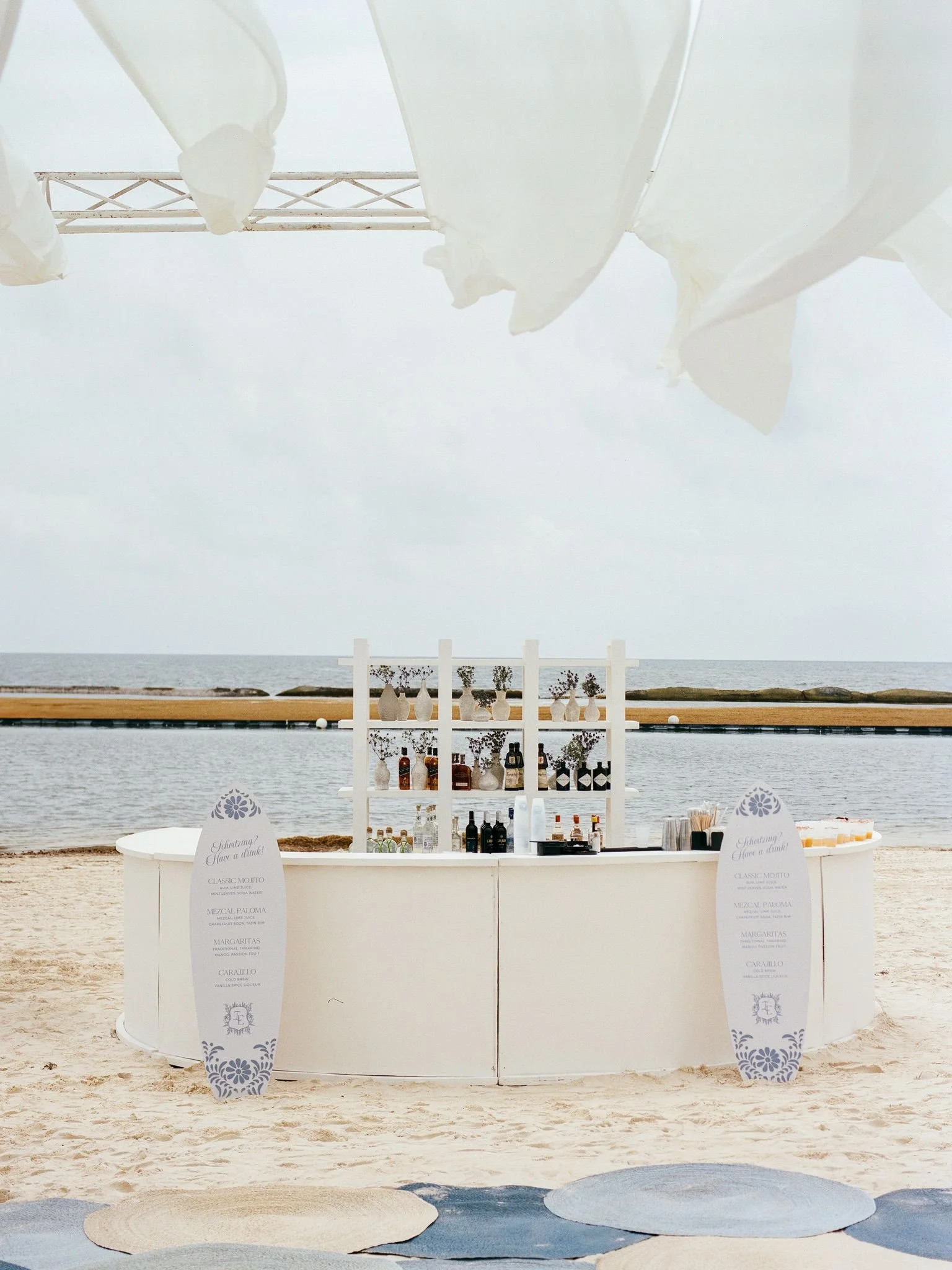 A beachside bar setup with bottles and glasses, decorated with vases and flowers, under white fabric canopies, against a backdrop of the ocean and sky.