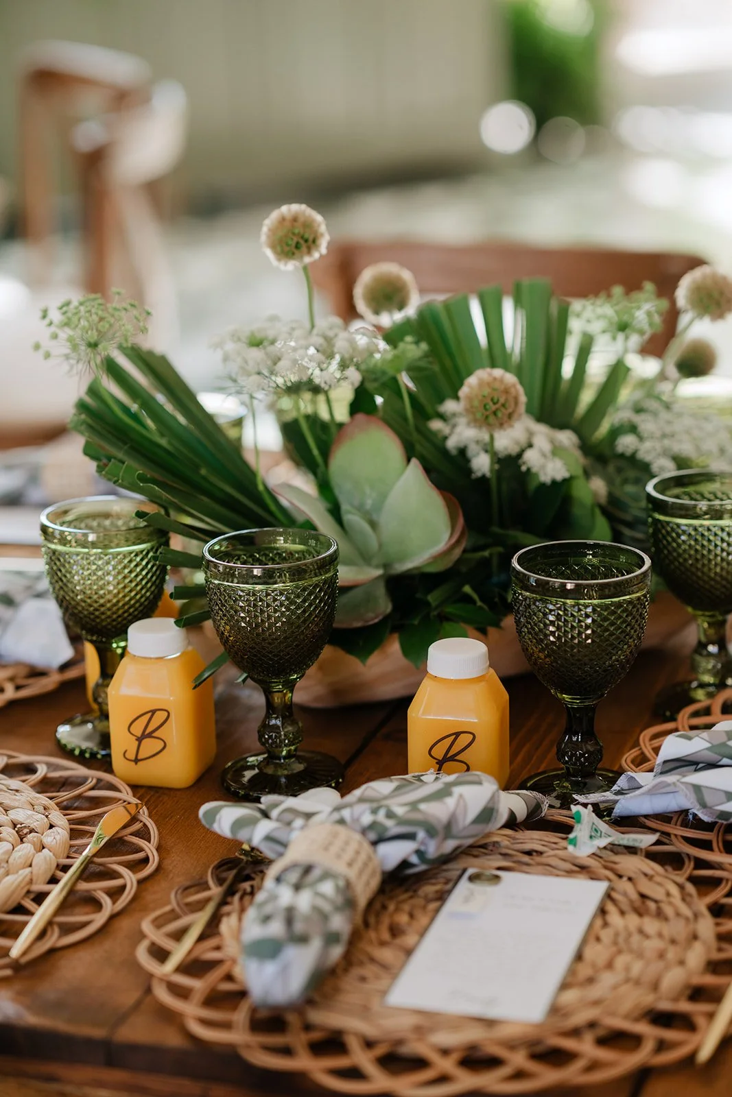 Table centerpiece with greenery and white flowers, surrounded by green glass goblets, yellow juice bottles, and woven placemats on a wooden table.