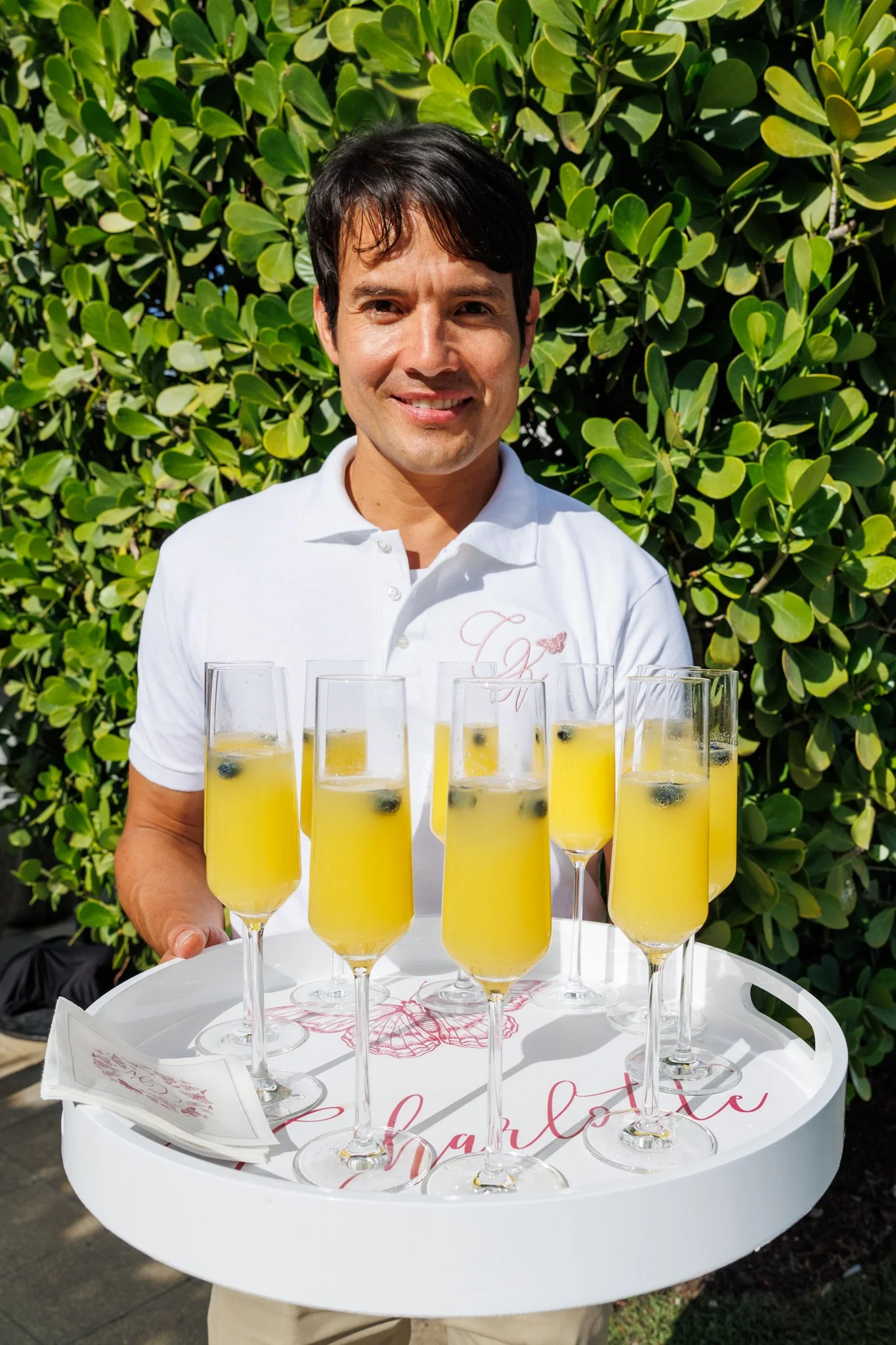 A man wearing a white polo shirt holding a tray with multiple glasses of yellow drink garnished with blueberries, standing outdoors in front of green foliage.