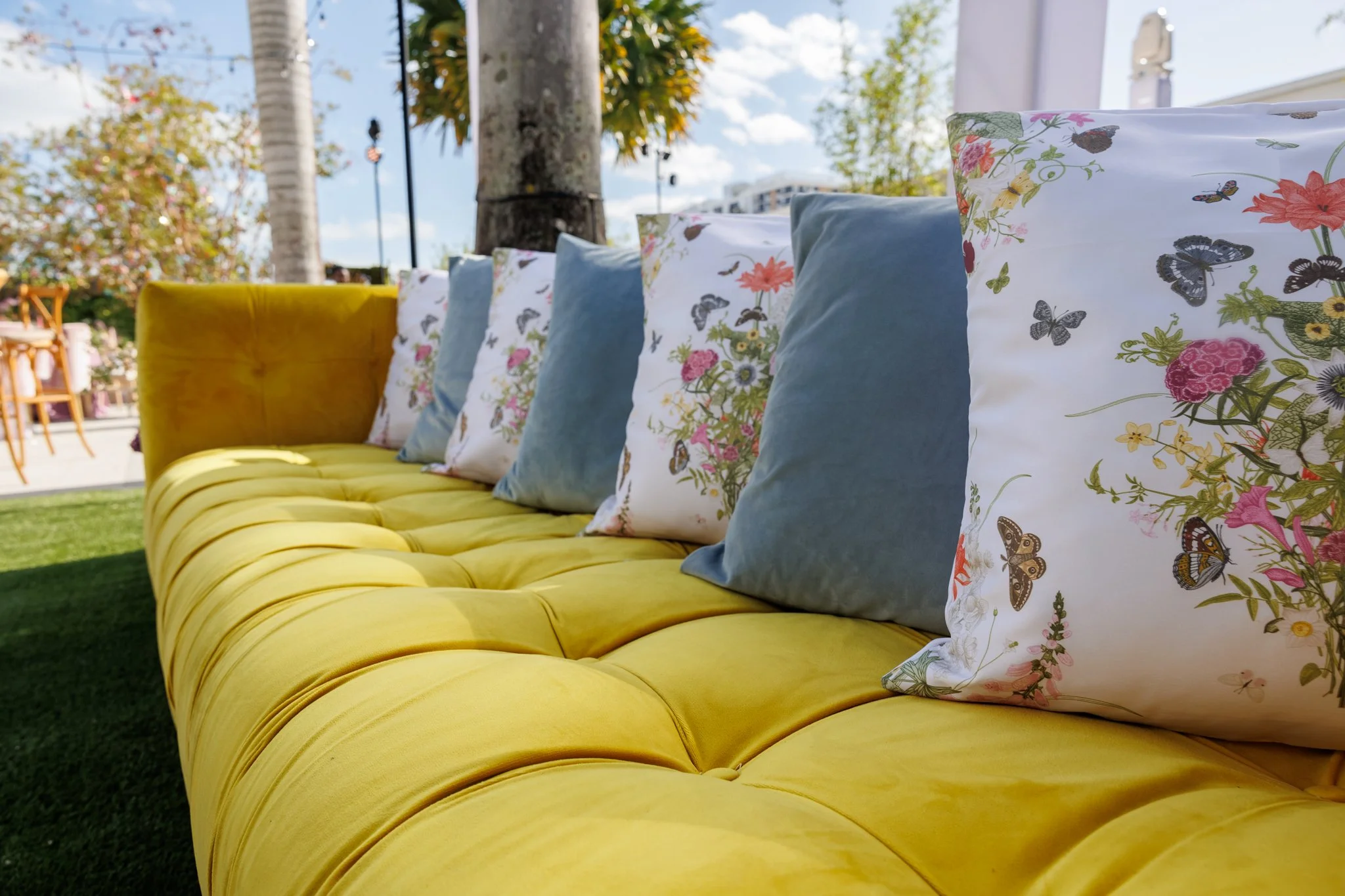 A yellow cushioned outdoor sofa with a row of decorative pillows featuring floral and butterfly patterns, set against a background of trees and a clear blue sky.