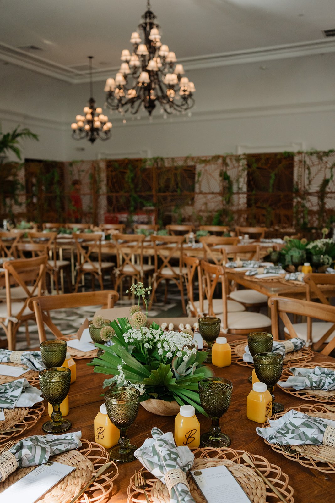 Elegant indoor event space decorated with wooden tables and chairs, featuring floral centerpieces, green glassware, and juice bottles, with chandeliers hanging from the ceiling and a backdrop with greenery.