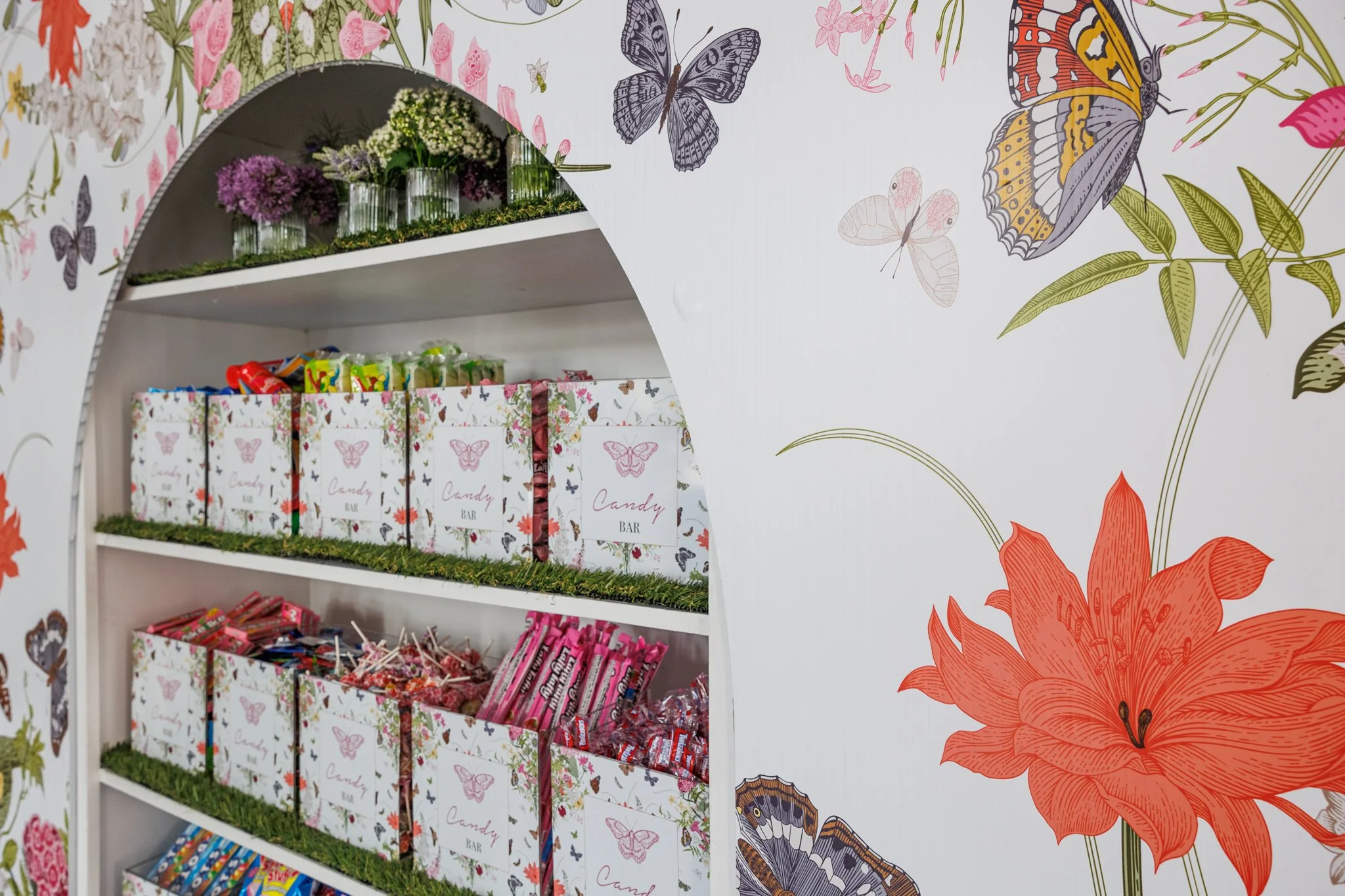 A display shelf with candies and snack packages against a wall decorated with colorful butterfly and floral wallpaper.