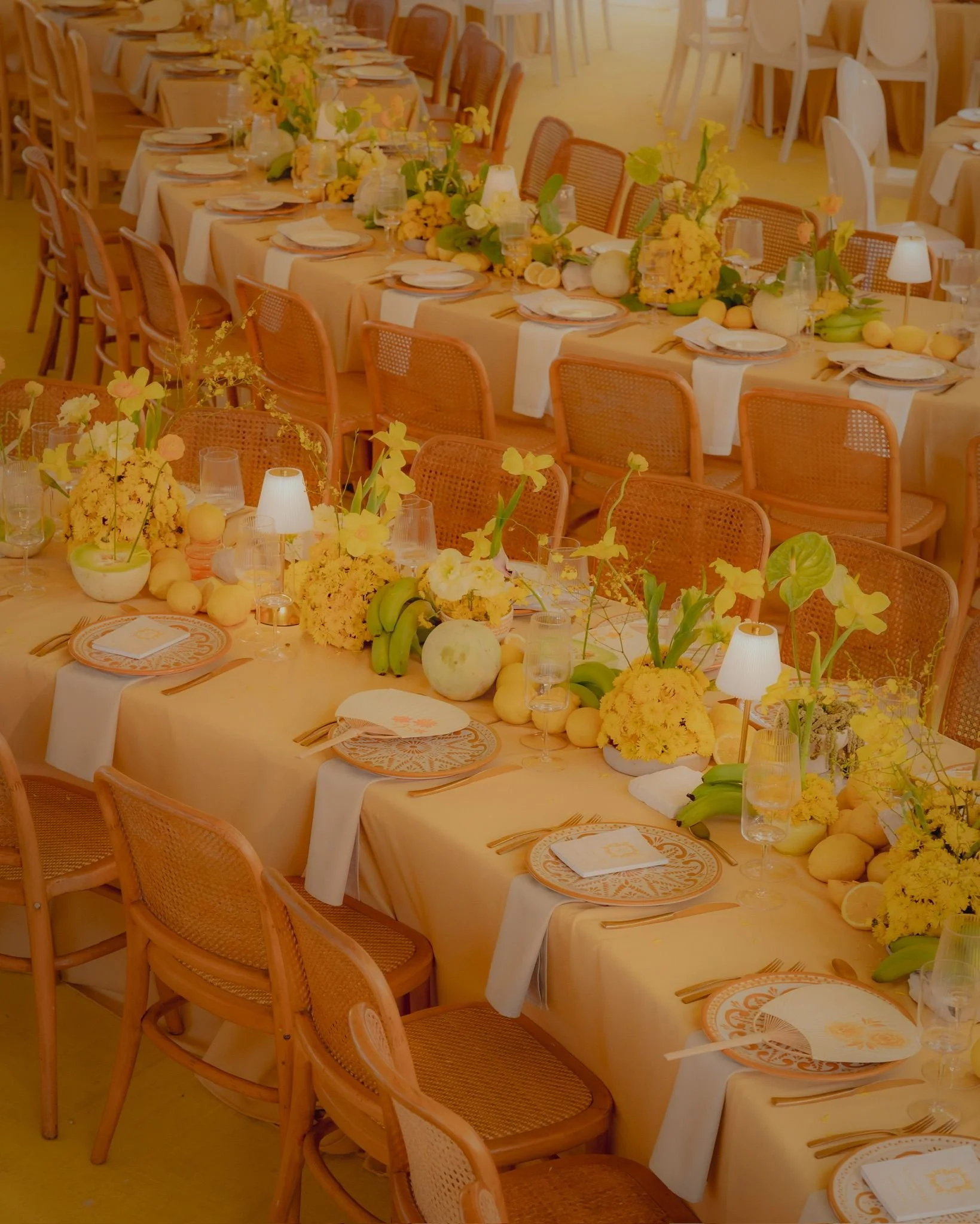 Decorated banquet table with yellow and orange accents, floral centerpieces, and place settings, in a well-lit indoor venue.