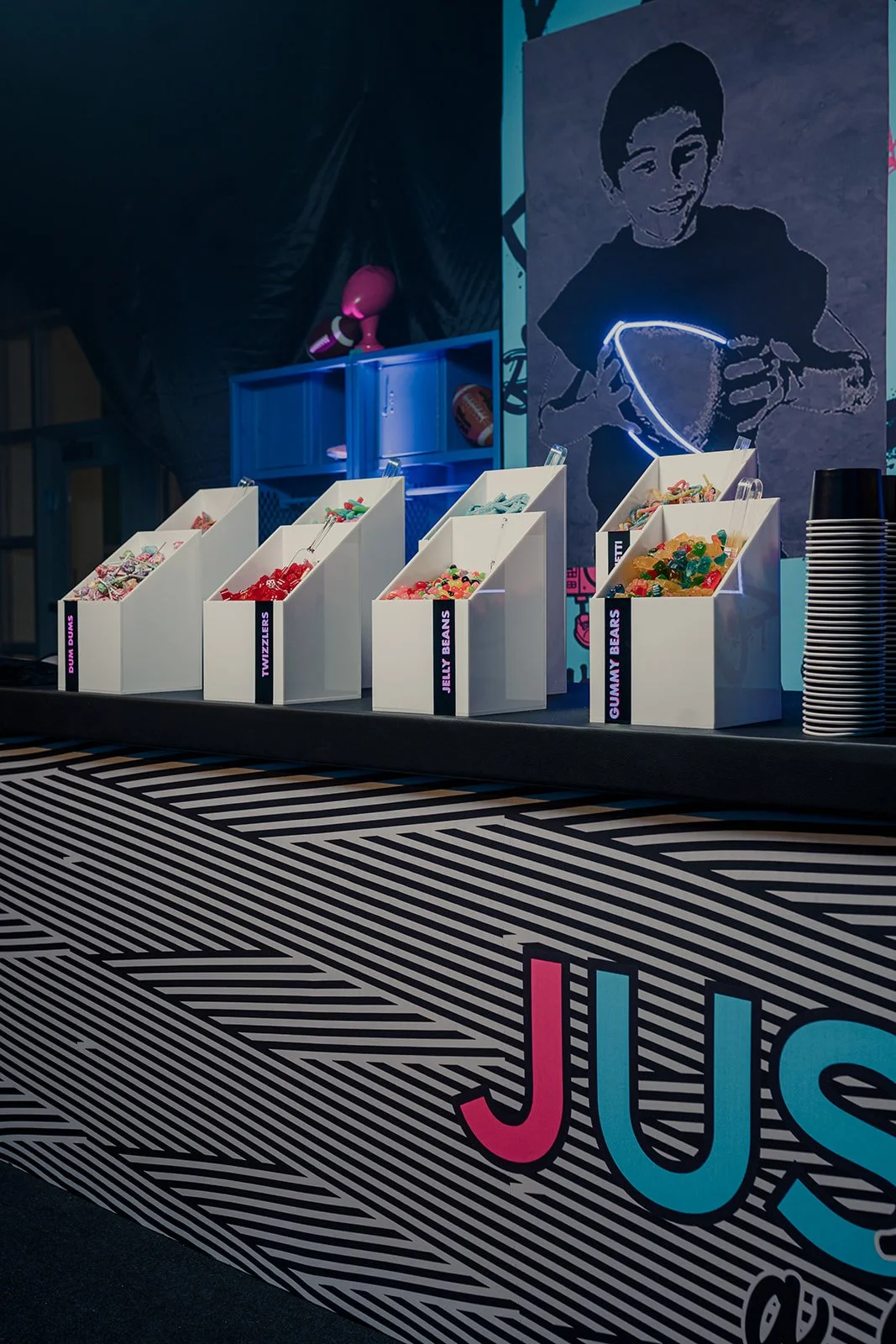 A candy buffet with various colorful candies organized in white bins labeled 'Dumb Dumbs,' 'Twizzlers,' 'Jelly Beans,' and 'Gummy Bears,' set against a black and white striped tablecloth with large colorful letters spelling 'JUS.' In the background, 