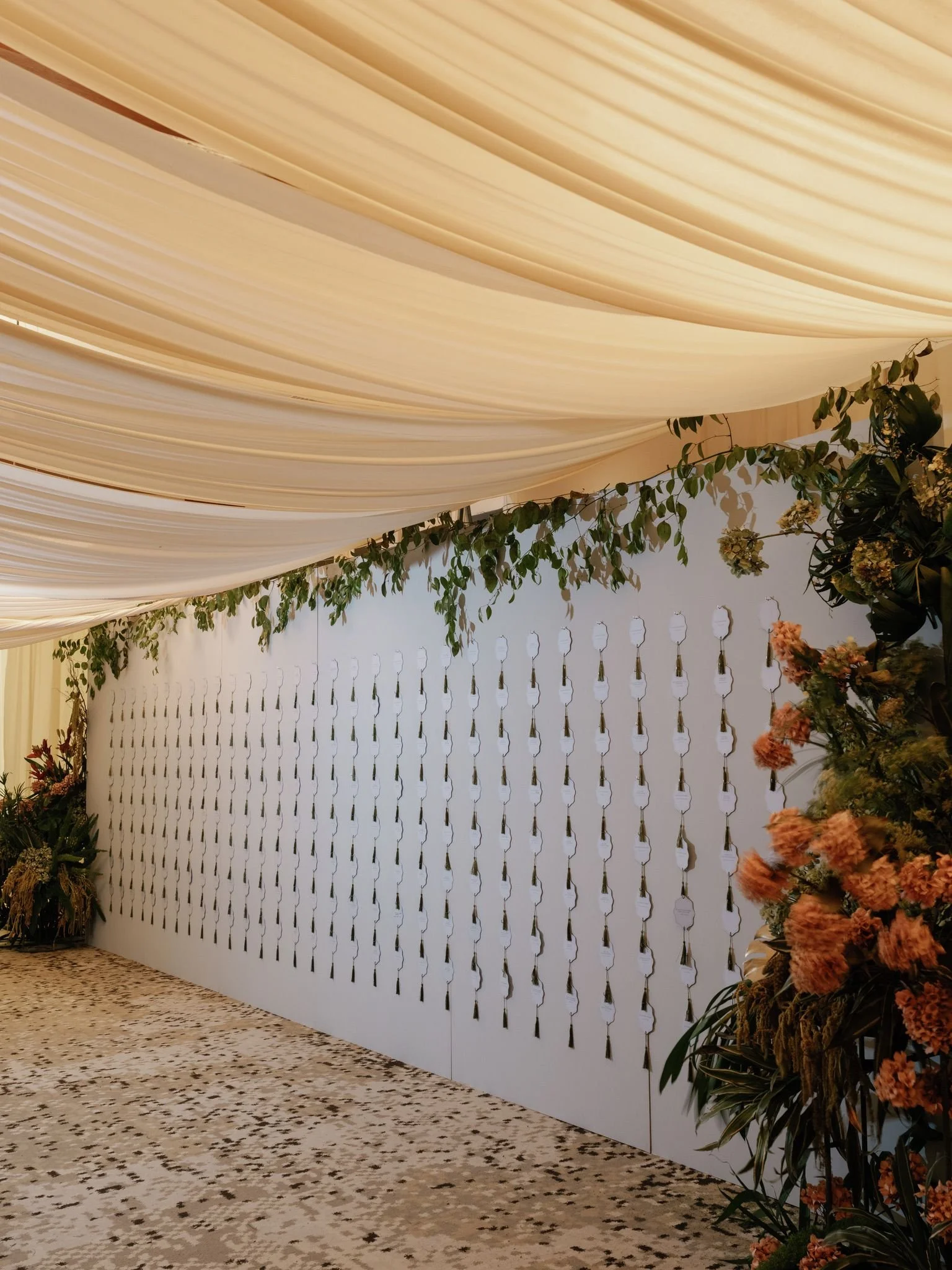 Decorative backdrop with cream-colored draped fabric ceiling, white wall with hanging tags or cards, and floral arrangements with green leaves and peach-colored flowers on the right side.