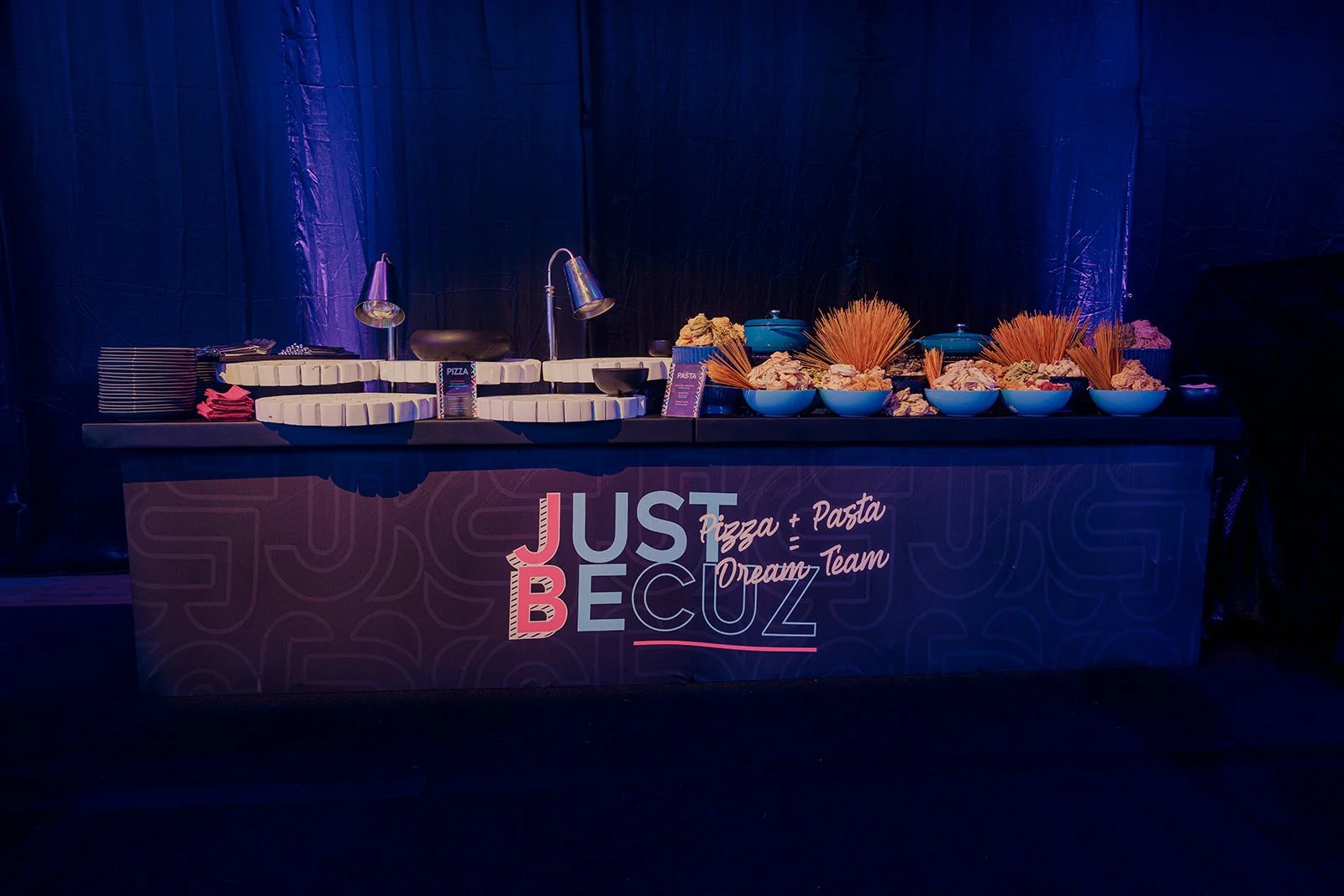 Buffet table with plates, bowls of pasta, and food, with a sign that says "Just Be You," in a dark setting with purple lighting.