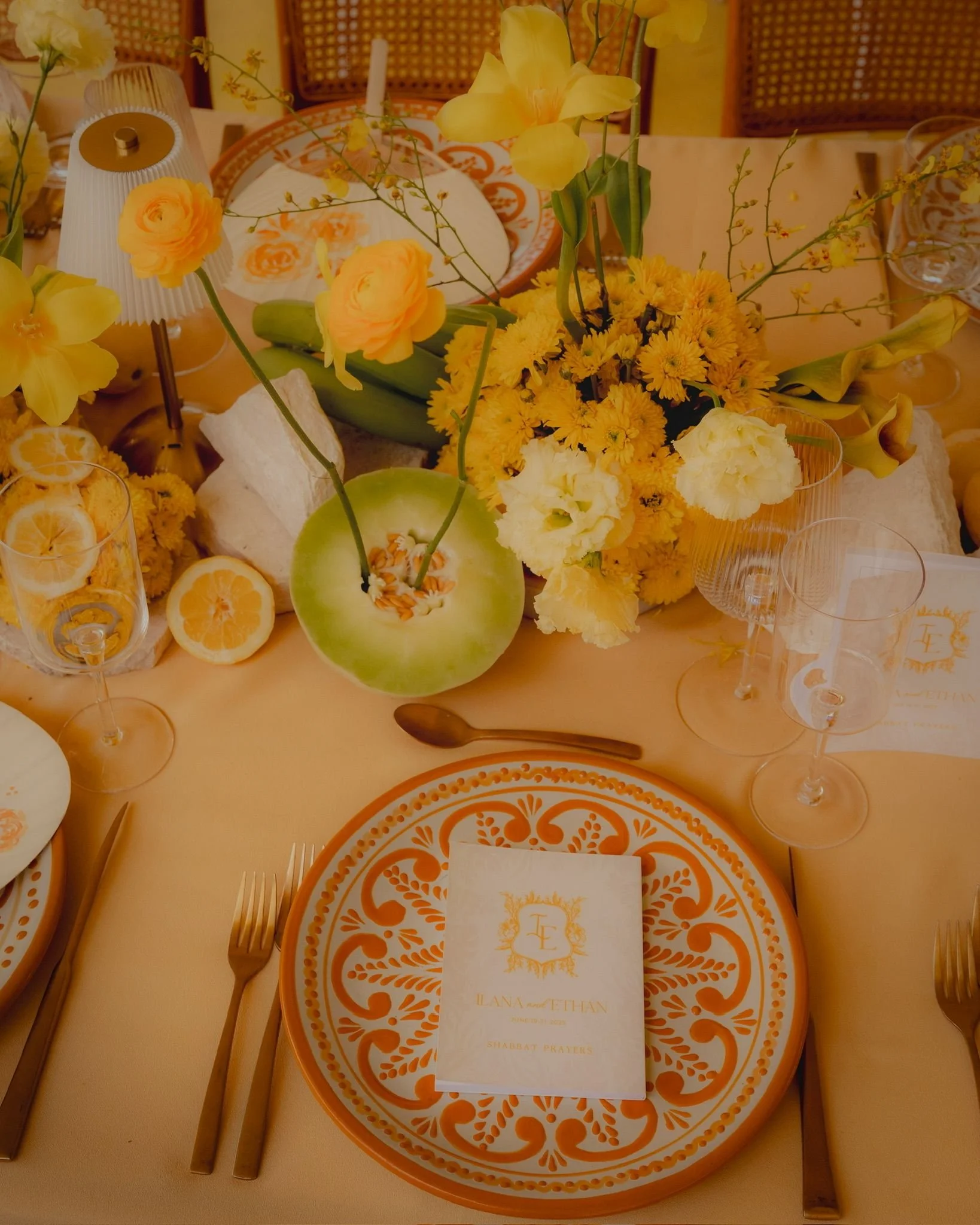 A decorative table setting with a floral centerpiece consisting of yellow flowers, including orchids and daisies, on a table with patterned orange and white plates, clear glassware, and a white napkin with a gold emblem. The table appears to be prepa
