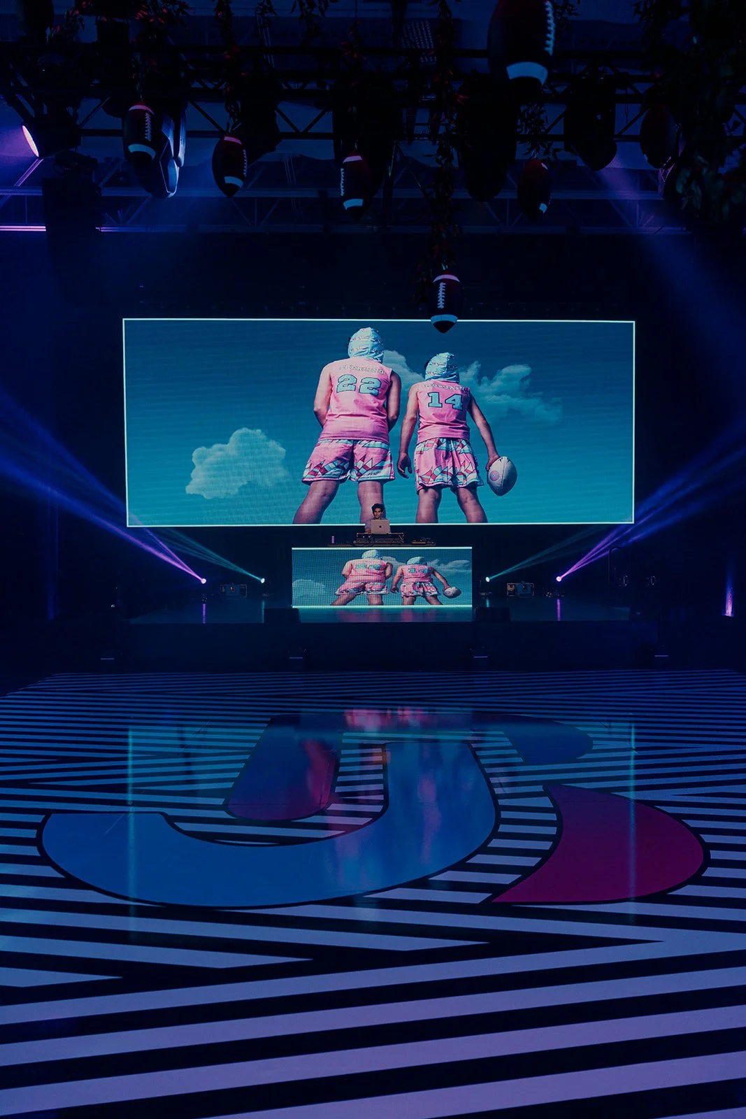 A stage with two large screens displaying images of two women in colorful sports outfits holding a football and volleyball, with a blue sky and clouds background. The stage has purple and blue lighting and a black and white striped floor with a large
