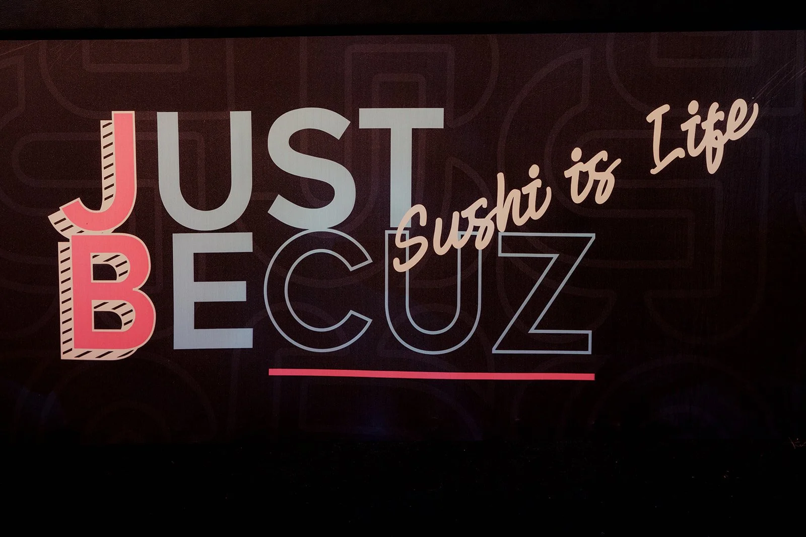 Neon sign with the text 'Just Be' in large letters, and 'Sushi is Life' written in cursive, with a dark background.