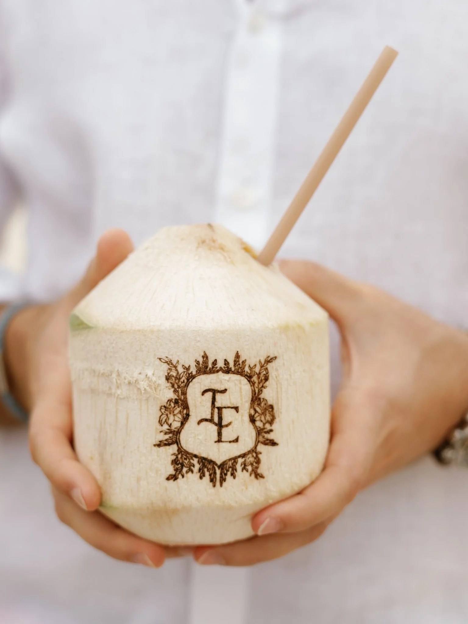 A person holding a young coconut with a straw inserted for drinking, the coconut has an engraved decorative design with the initials 'T F'.