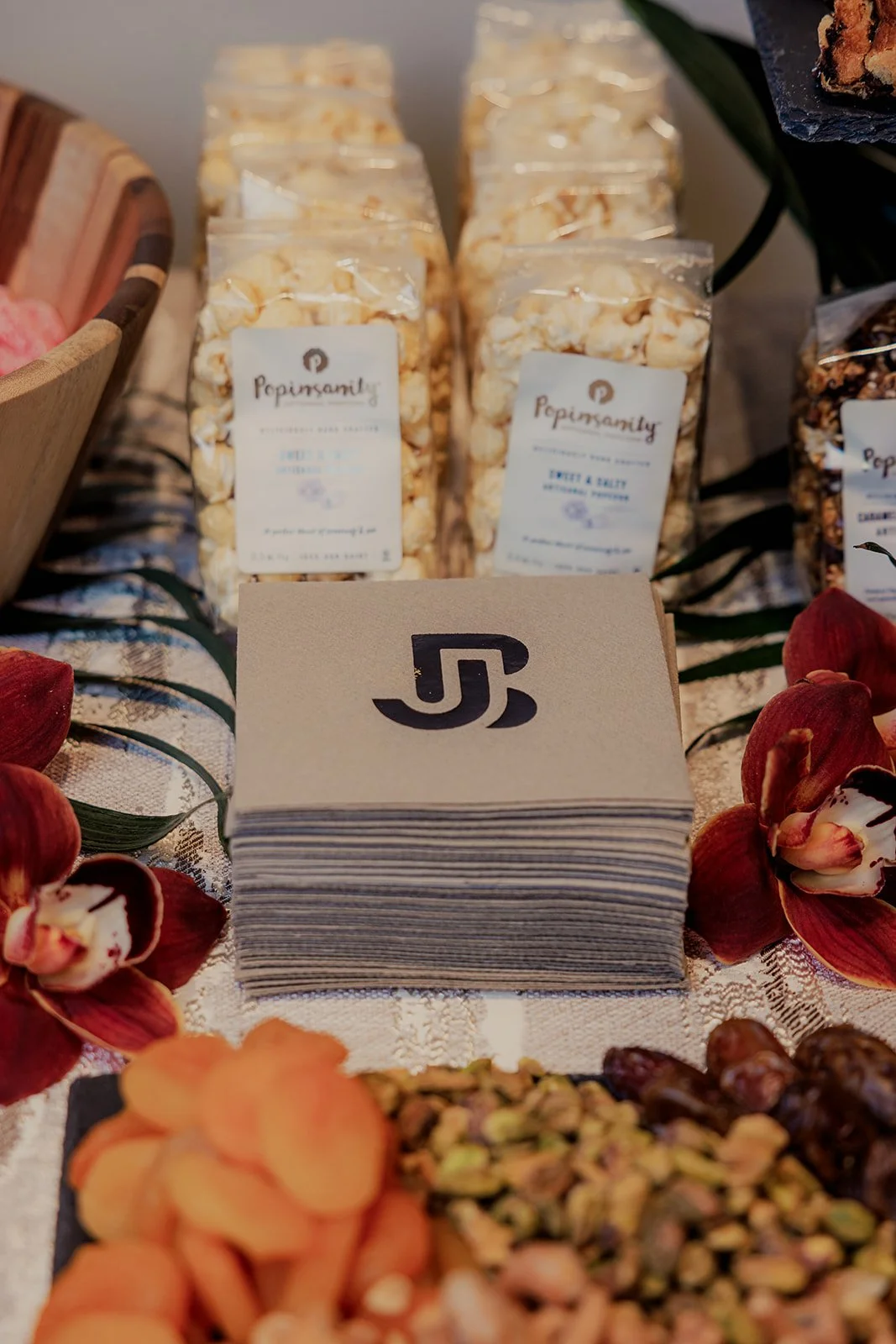 Small glass jars of popcorn labeled 'Popinsanity' on a table, decorated with flower petals, and a stack of branded napkins in the foreground.