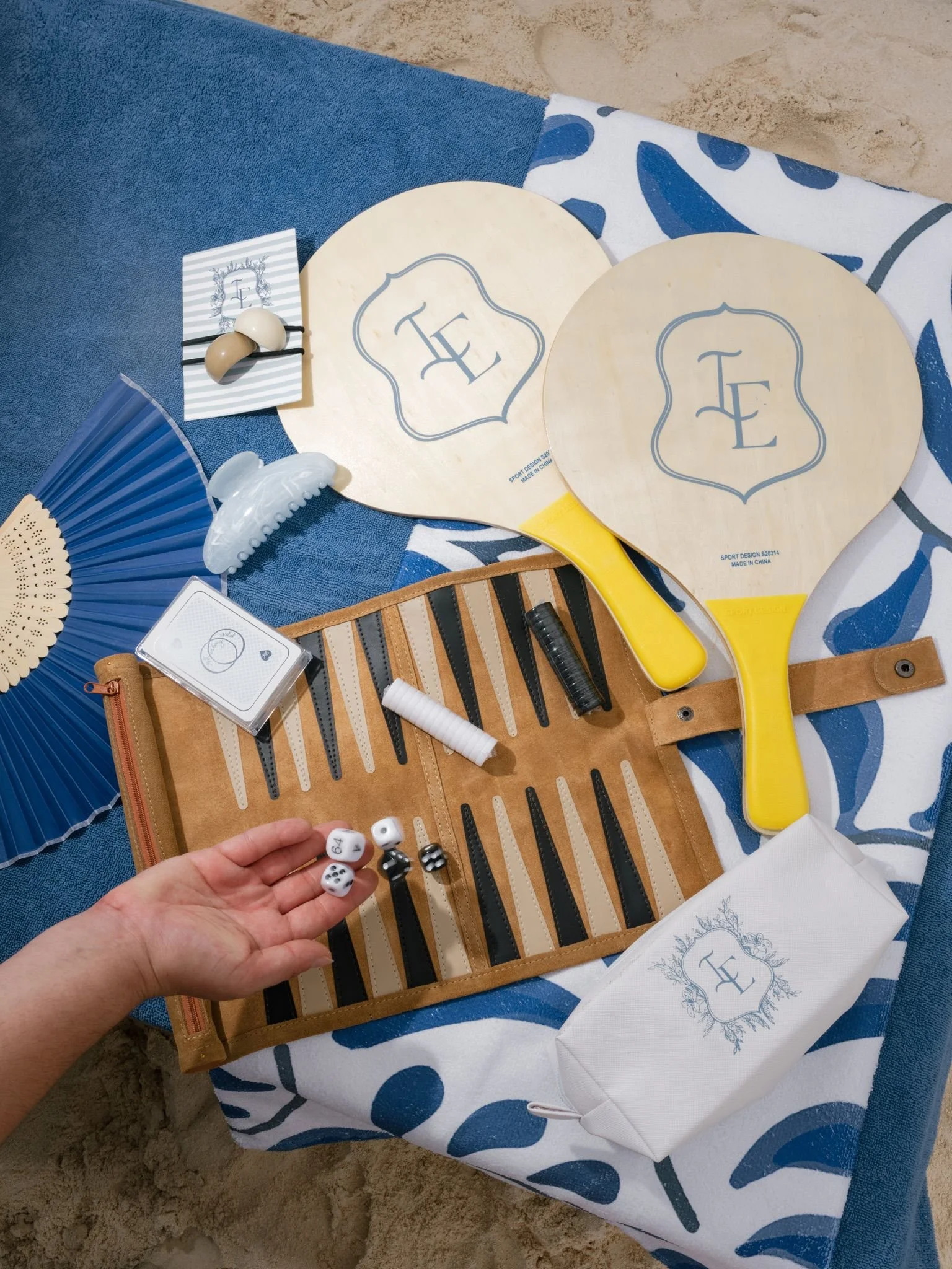 Beach scene with a table covered in a patterned blue and white cloth, holding a paddle, decorative items, and monogrammed towels. Sand is visible on the ground.
