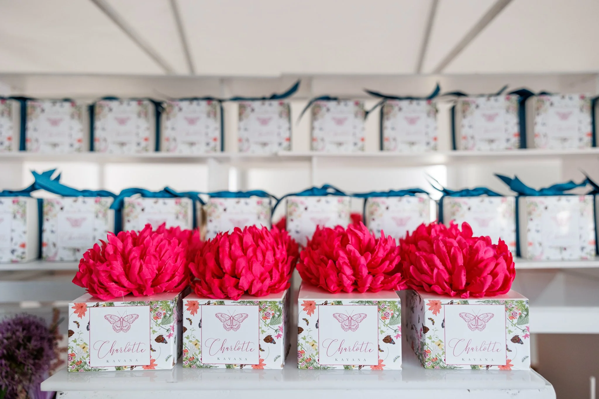 Bright pink flower arrangements in decorative boxes labeled 'Charlotte Kavana' with butterfly and floral designs, on a white display shelf.