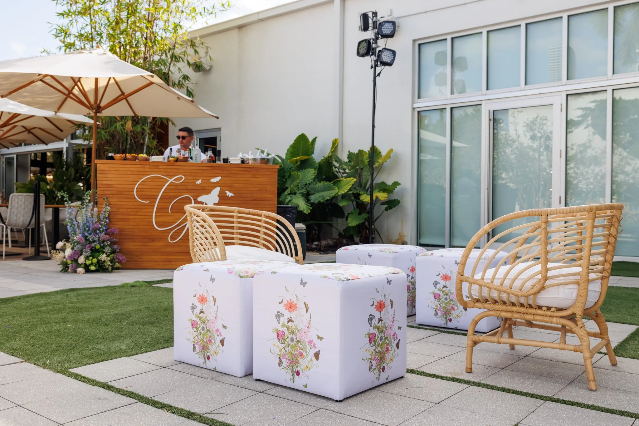 Outdoor patio with wicker chairs, floral-patterned ottomans, a bar with a man serving drinks, large umbrellas, greenery, and a modern building with large windows.