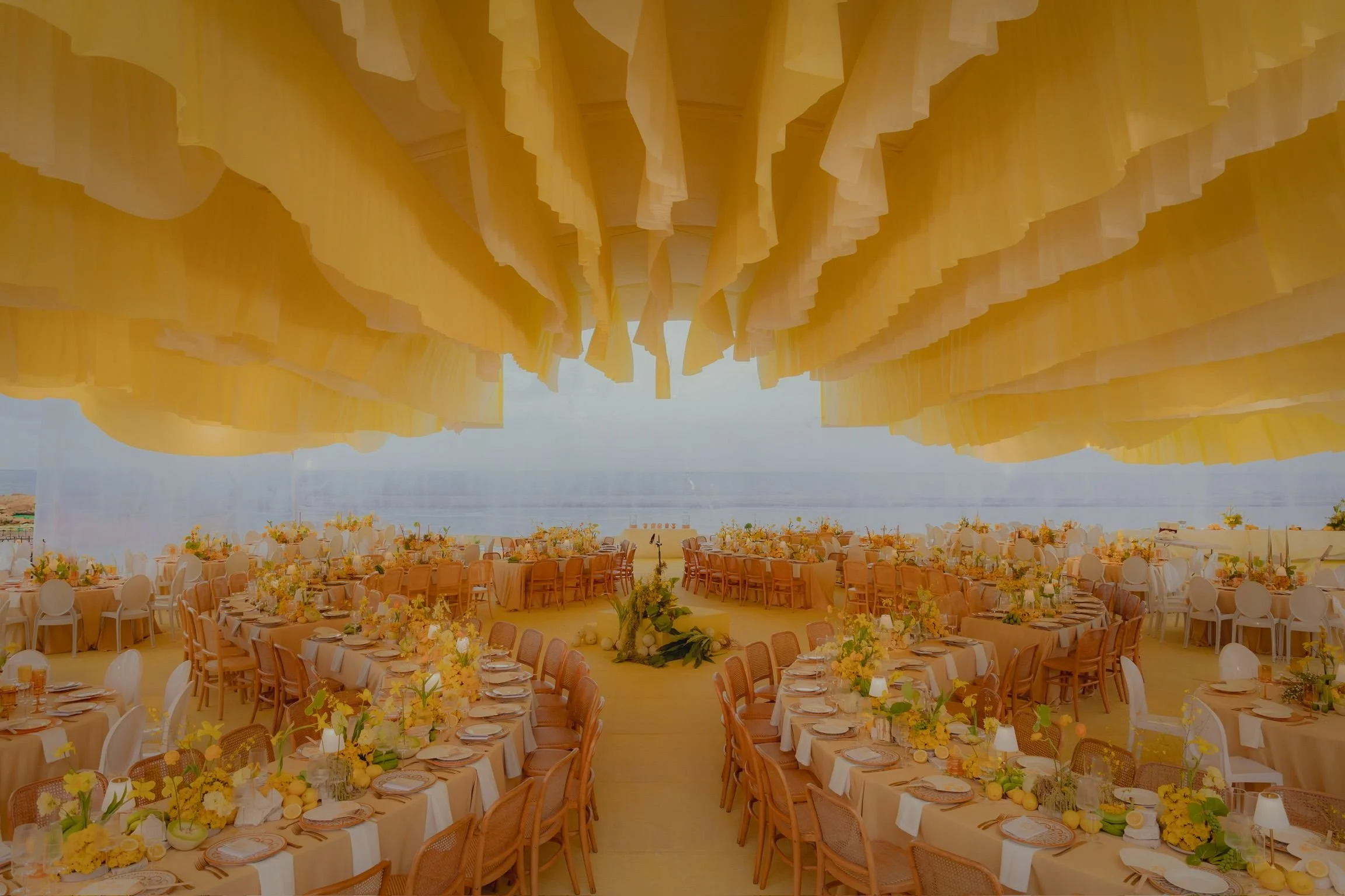 Indoor wedding reception setup under a large, yellow fabric canopy overlooking a body of water. Round tables decorated with yellow flowers and place settings fill the space.