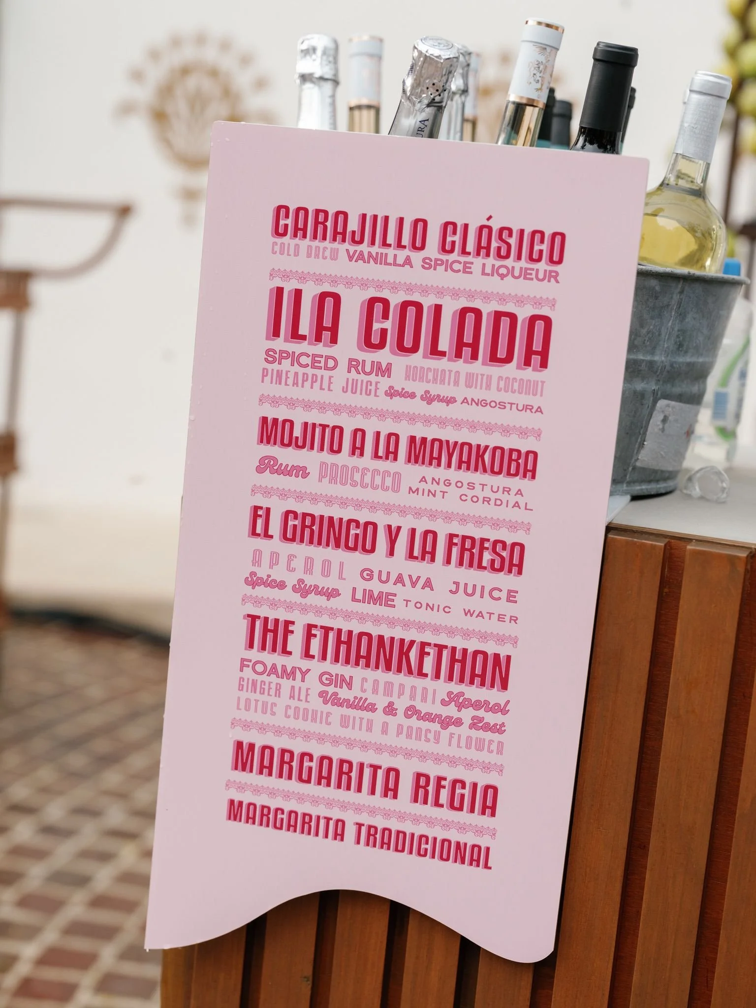 A pink menu displaying various cocktail names and ingredients stands on a wooden surface outside. Bottles of liquor are visible in the background.