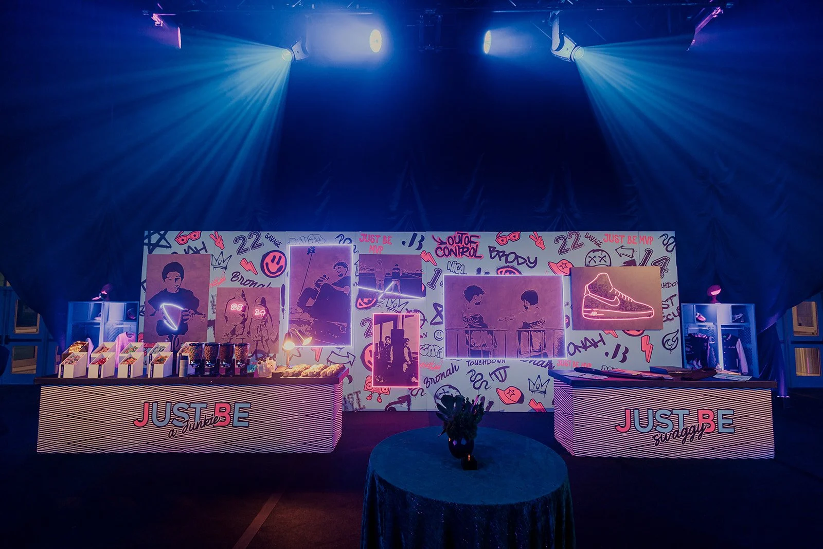 A decorated indoor event space with neon signs, graffiti-style wall art, and tables with party supplies. The backdrop features pink and purple lighting and cartoonish graffiti with phrases like 'Just Be' and 'Savage.'