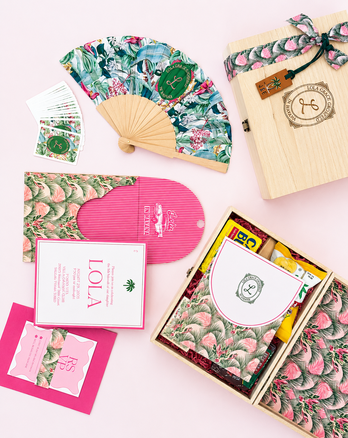A wooden gift box with a tropical leaf design on the cover, tied with a black strap and a small wooden tag, alongside a folding fan, a pack of playing cards, and various wrapped gifts on a pink background.