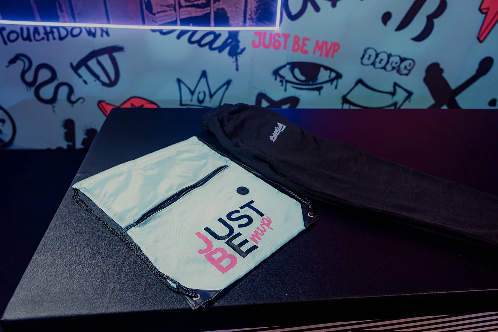 A white and black bag with the words "Just Be MVP" on it, placed on a black table. Behind it, there is a black clothing item, possibly a jacket or pants. The background features graffiti-style art with various doodles, an eye, the words "Touchdown" a