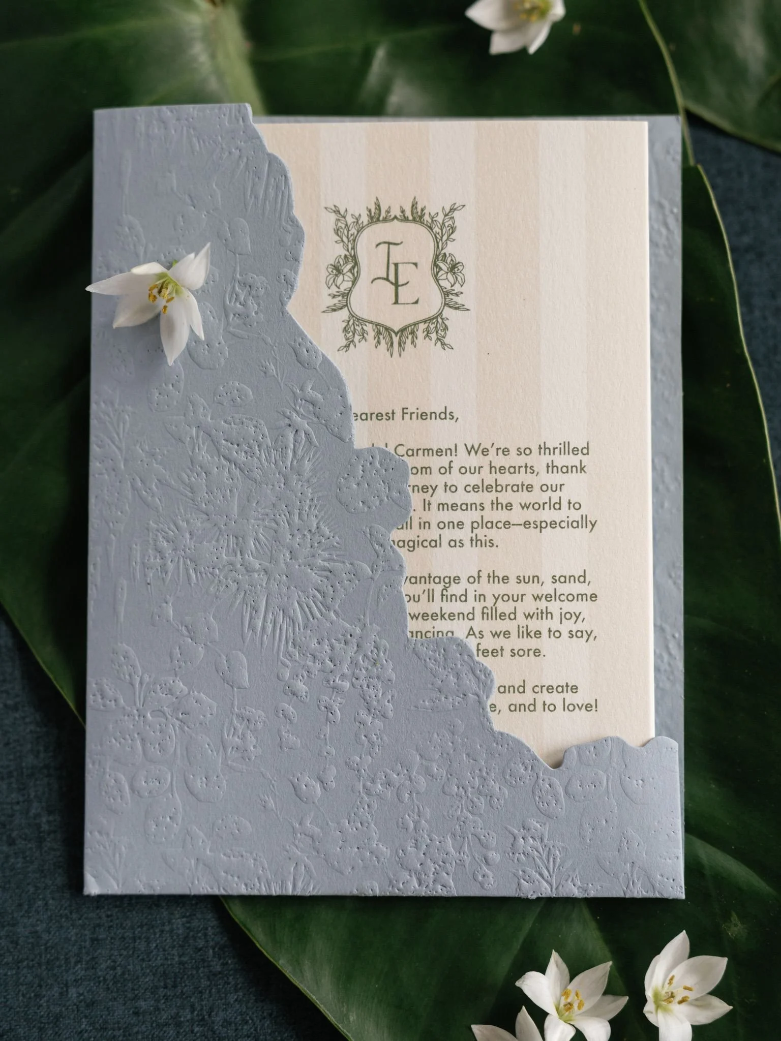 A wedding or event invitation with a monogram 'LE' in a decorative frame, placed on a white textured paper with embossed floral patterns. The invitation is partially covered by a white flower and is set on green leaves with additional white flowers a
