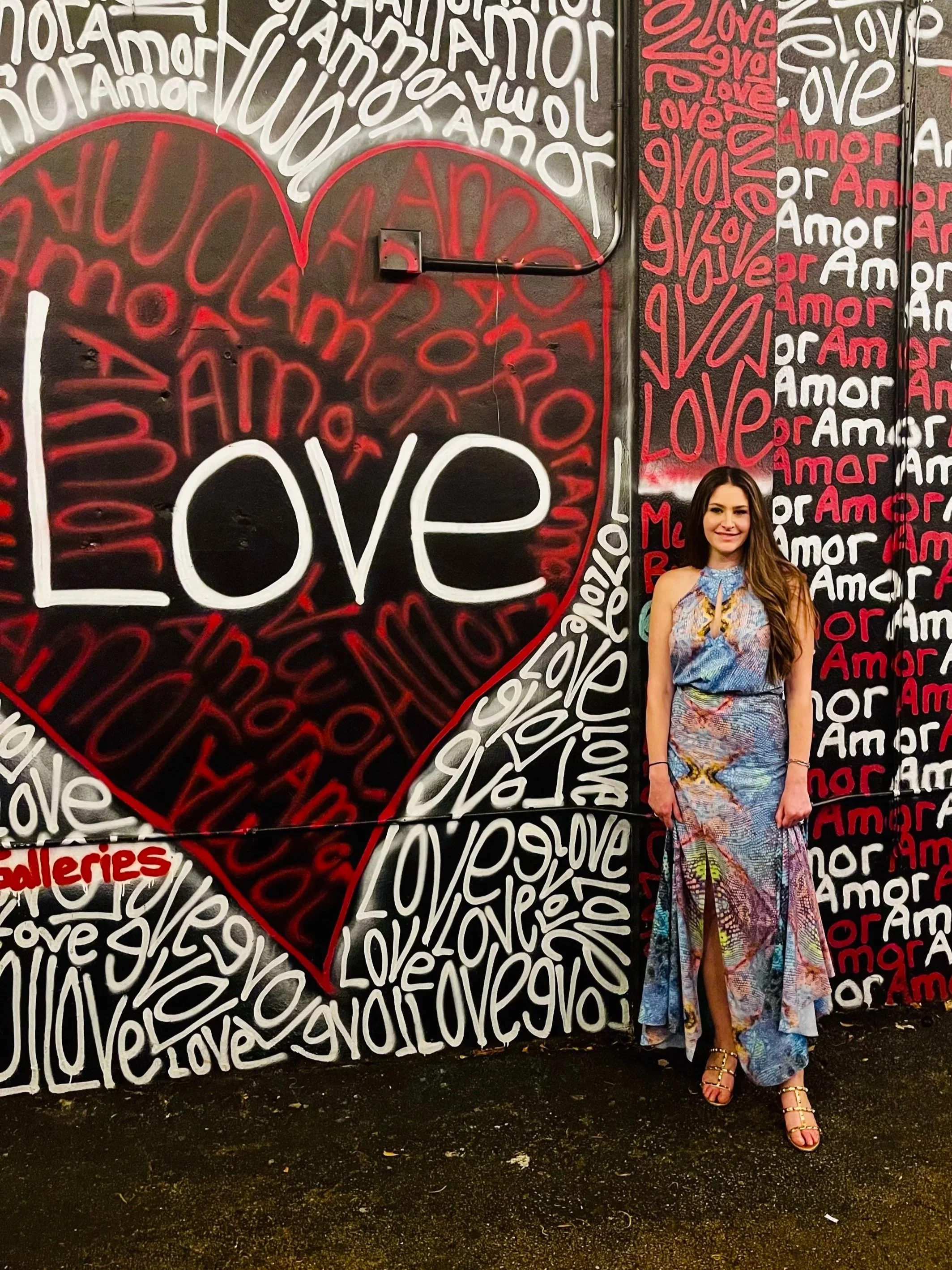 A woman standing in front of a colorful mural with the word 'LOVE' written repeatedly in various languages and colors.